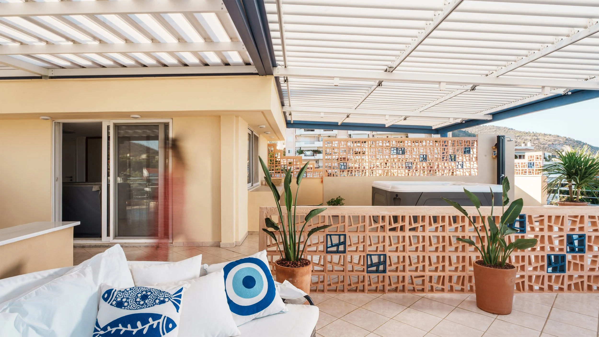 Balcony with white pergola roof, potted plants, outdoor hot tub, and sliding glass door leading to interior of a building with mountains in the background.