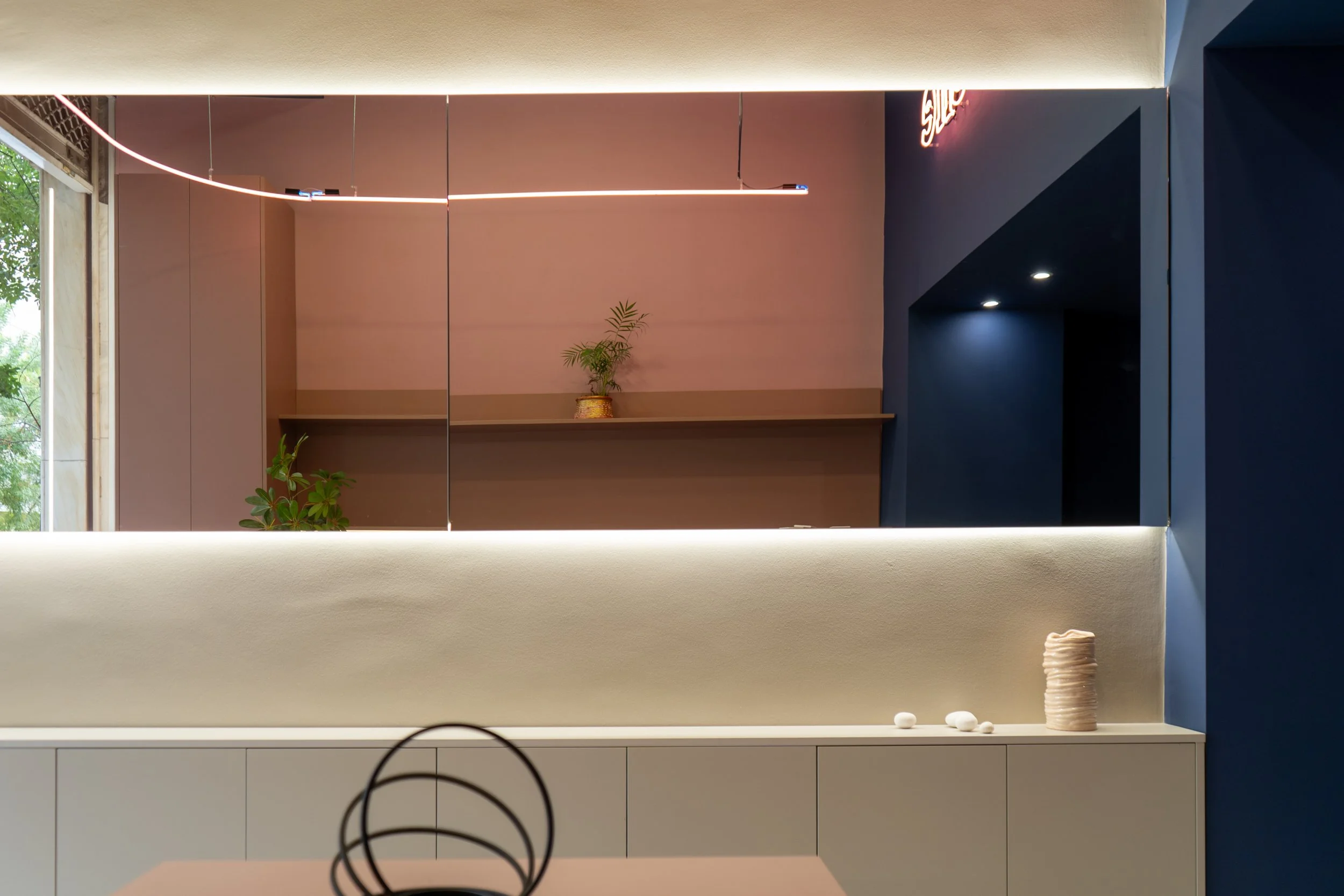 A modern interior space with a large mirror on the wall, providing a reflection of a pink wall with a small shelf holding a potted plant. The space features soft lighting and minimalist decor, including a decorative white vase and small white objects