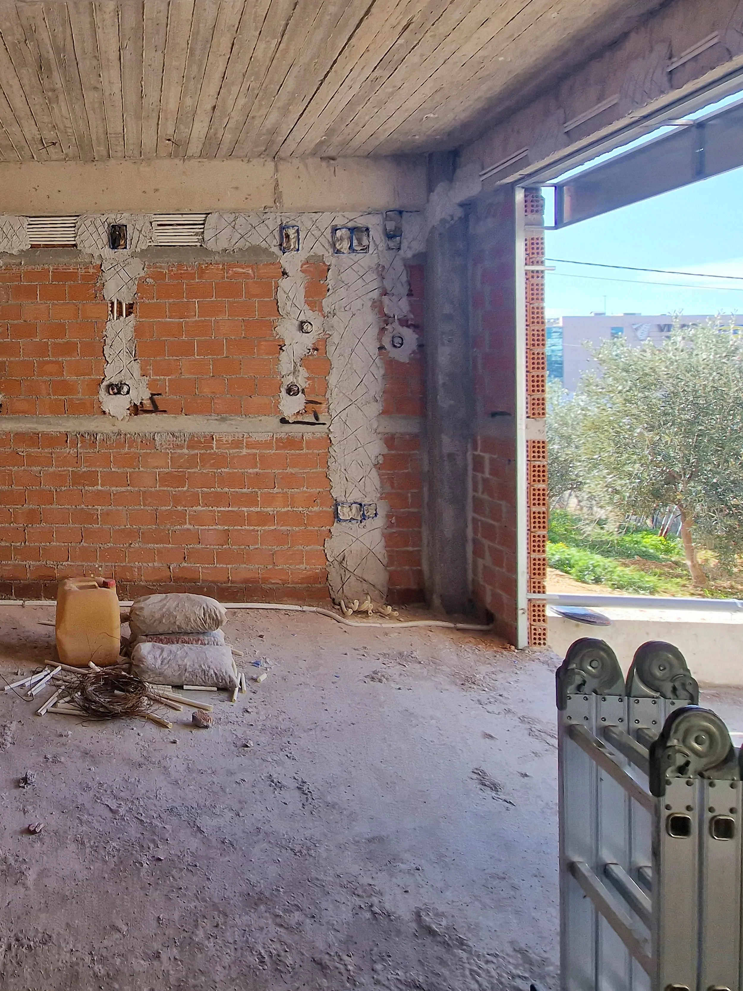 Interior view of a building under construction with brick walls, electrical wiring, construction materials on the floor, and a partially finished doorway opening to the outside where trees and buildings are visible.