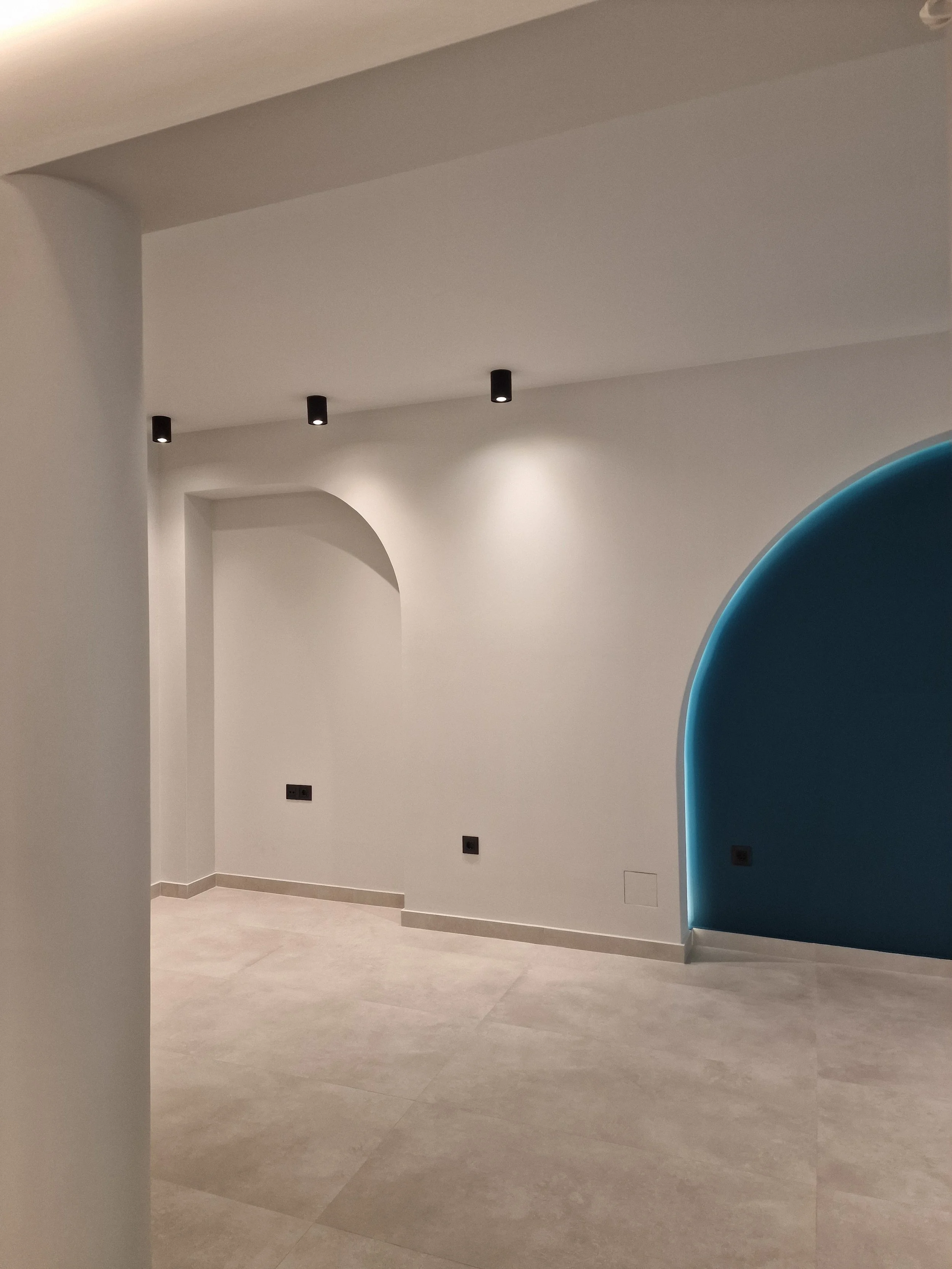 Empty interior space with white walls, beige tiled flooring, and a blue curved architectural feature on the right side. Ceiling lights are visible, and wall outlets are seen near the floor.