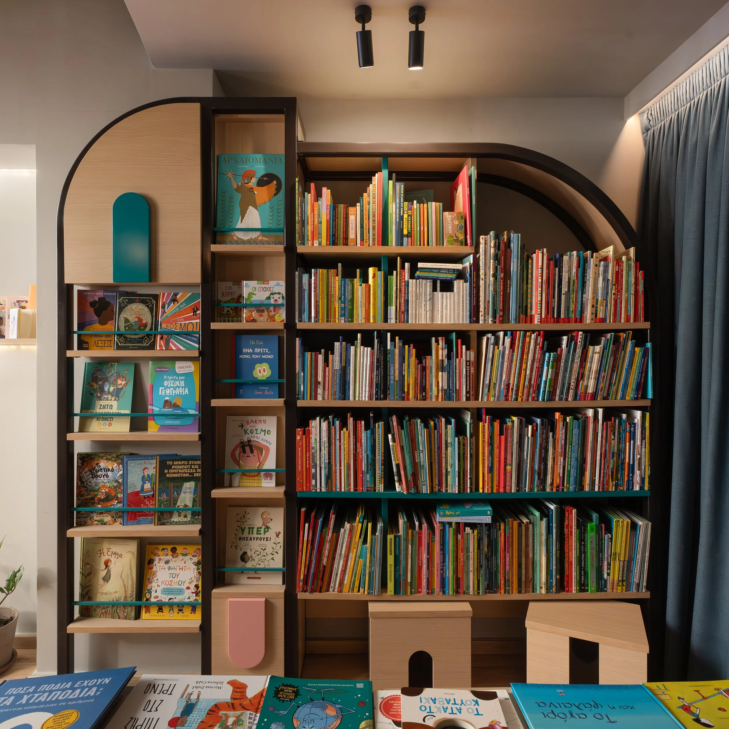 Bookshelves with colorful children's books and educational materials in a room with a modern interior, featuring a light gray ceiling with two black ceiling lights, gray curtains on the right, and wooden furniture.