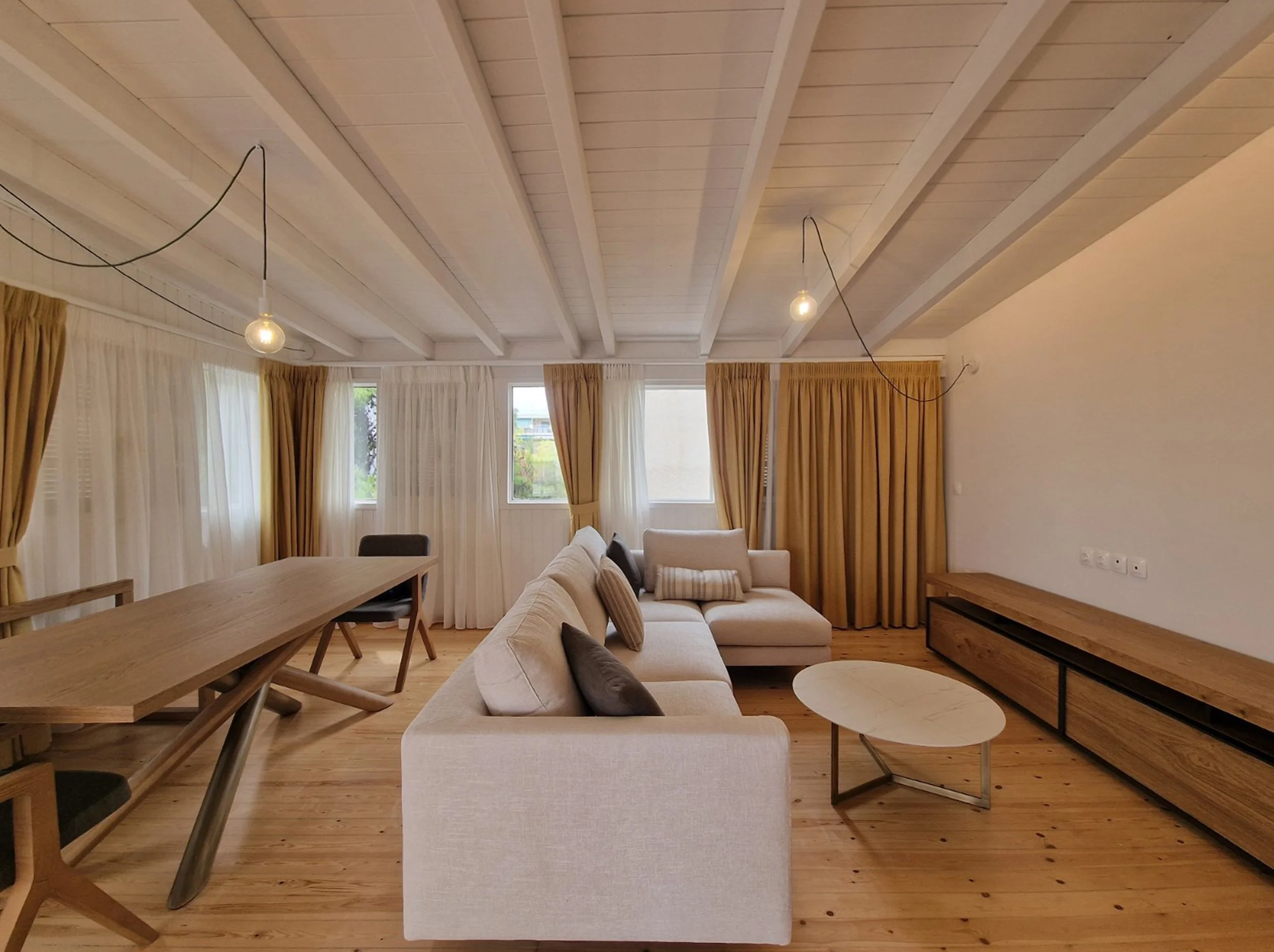 Living room with a beige sofa, wooden dining table and chairs, light wood floors, large windows with white and gold curtains, and modern hanging light bulbs.