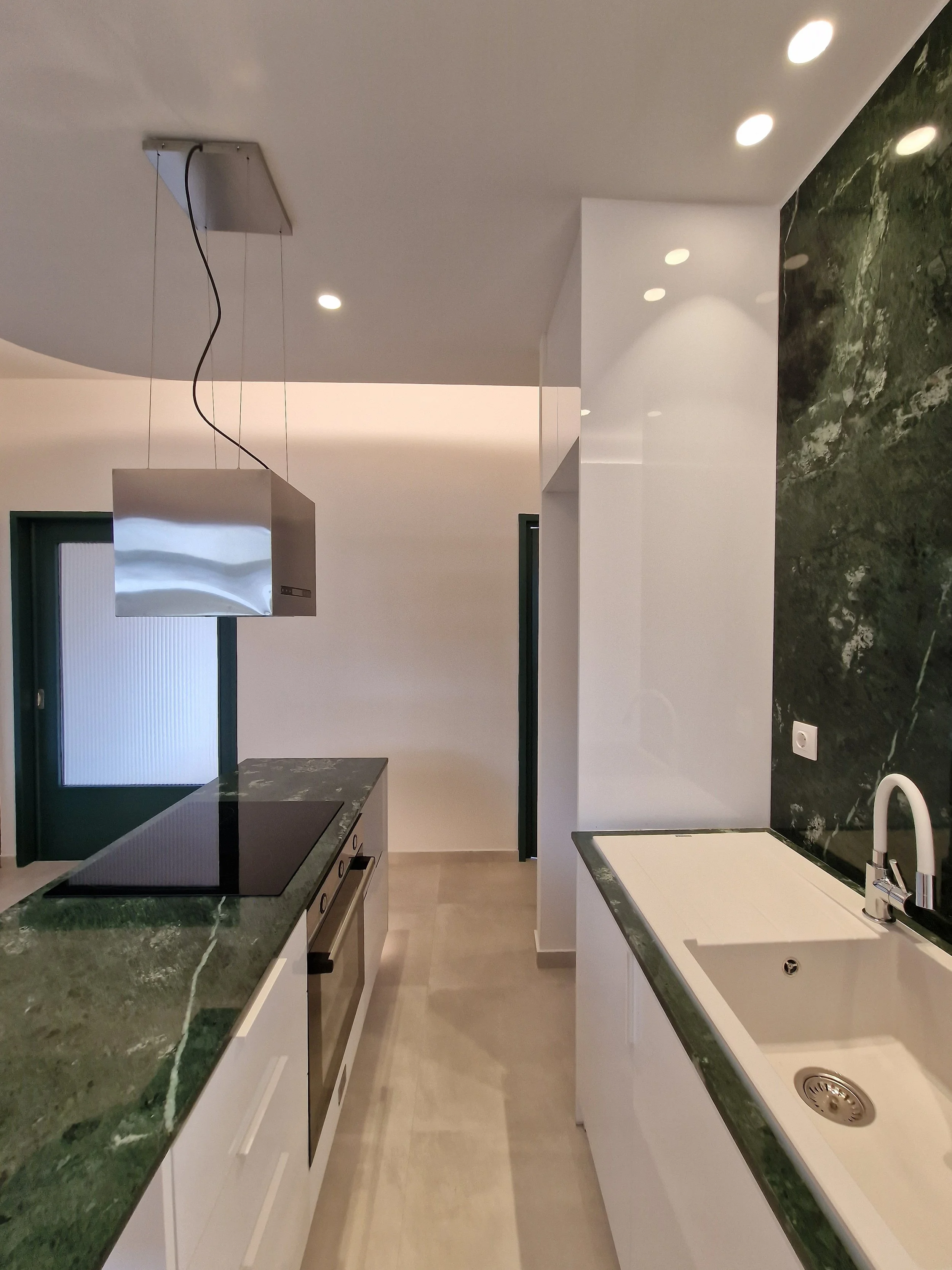 Modern kitchen with green marble countertops, white cabinetry, black electric cooktop, built-in oven, and white sink with a silver faucet. Recessed ceiling lights and partial view of a door.