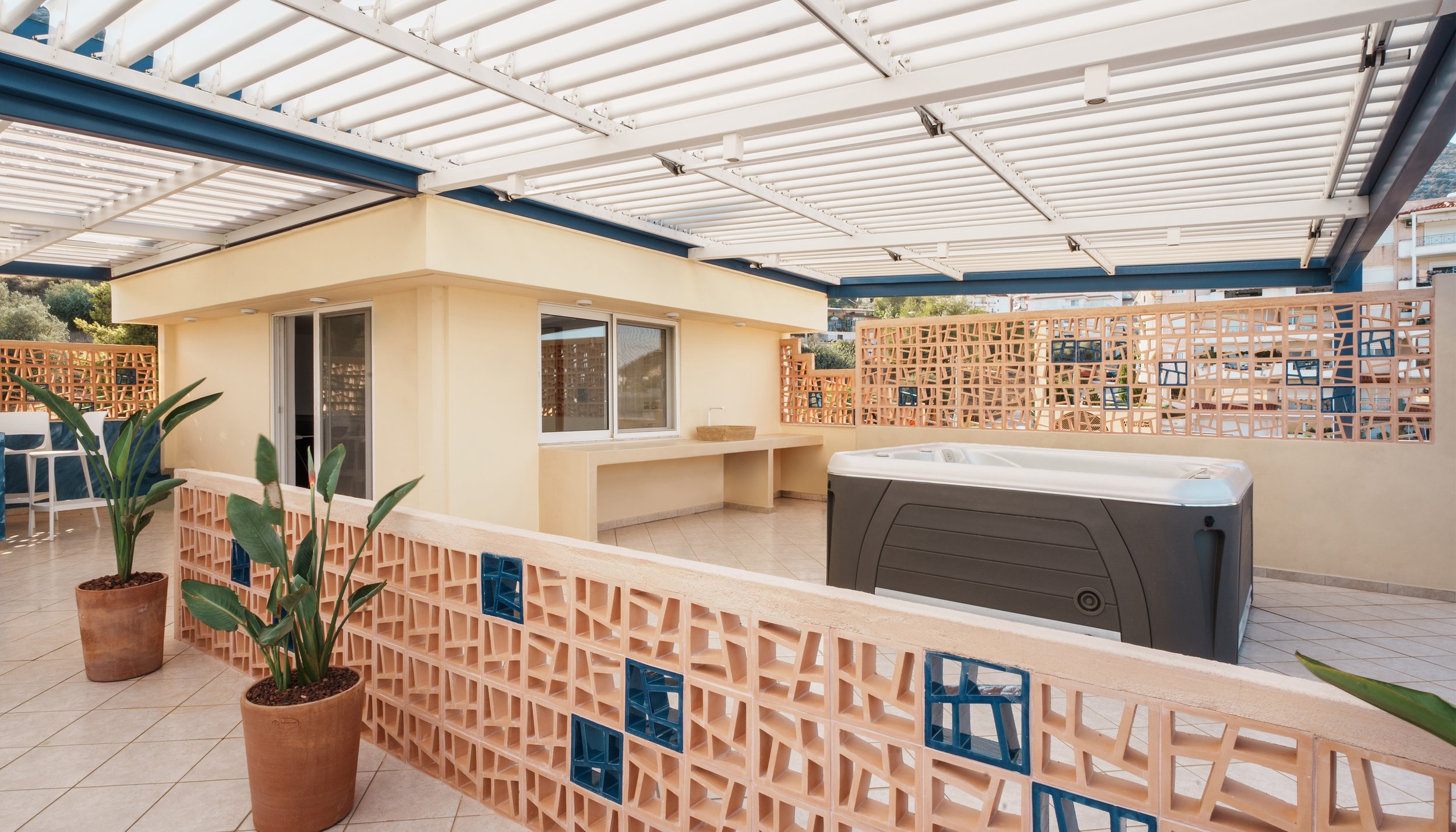 A covered outdoor terrace with potted plants, a beige wall, sliding glass door, and a hot tub, enclosed by a decorative brick wall with blue accents.