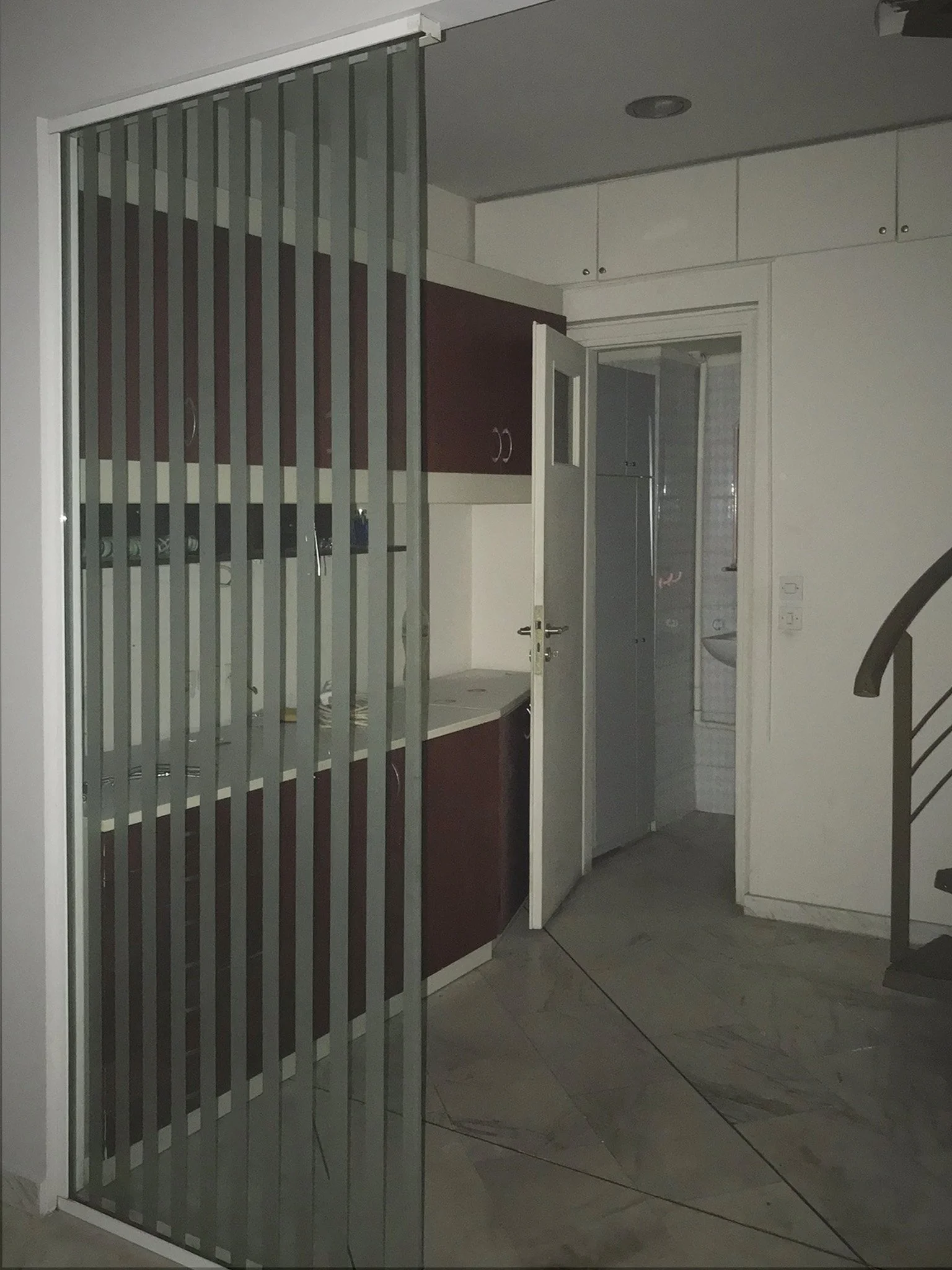 Interior of a room with gray tiled floor, a glass door with a metal security gate, wooden cabinets, and a partially open door leading to another room with white tiles.
