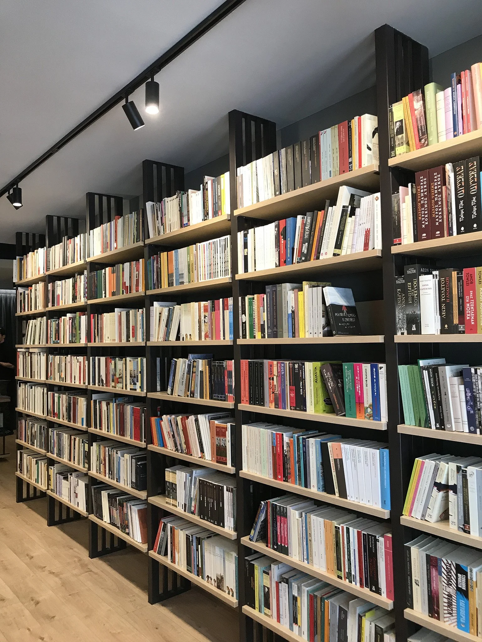 Bookshelves filled with books in a library or bookstore, with a light-colored wooden floor and ceiling-mounted black track lighting.