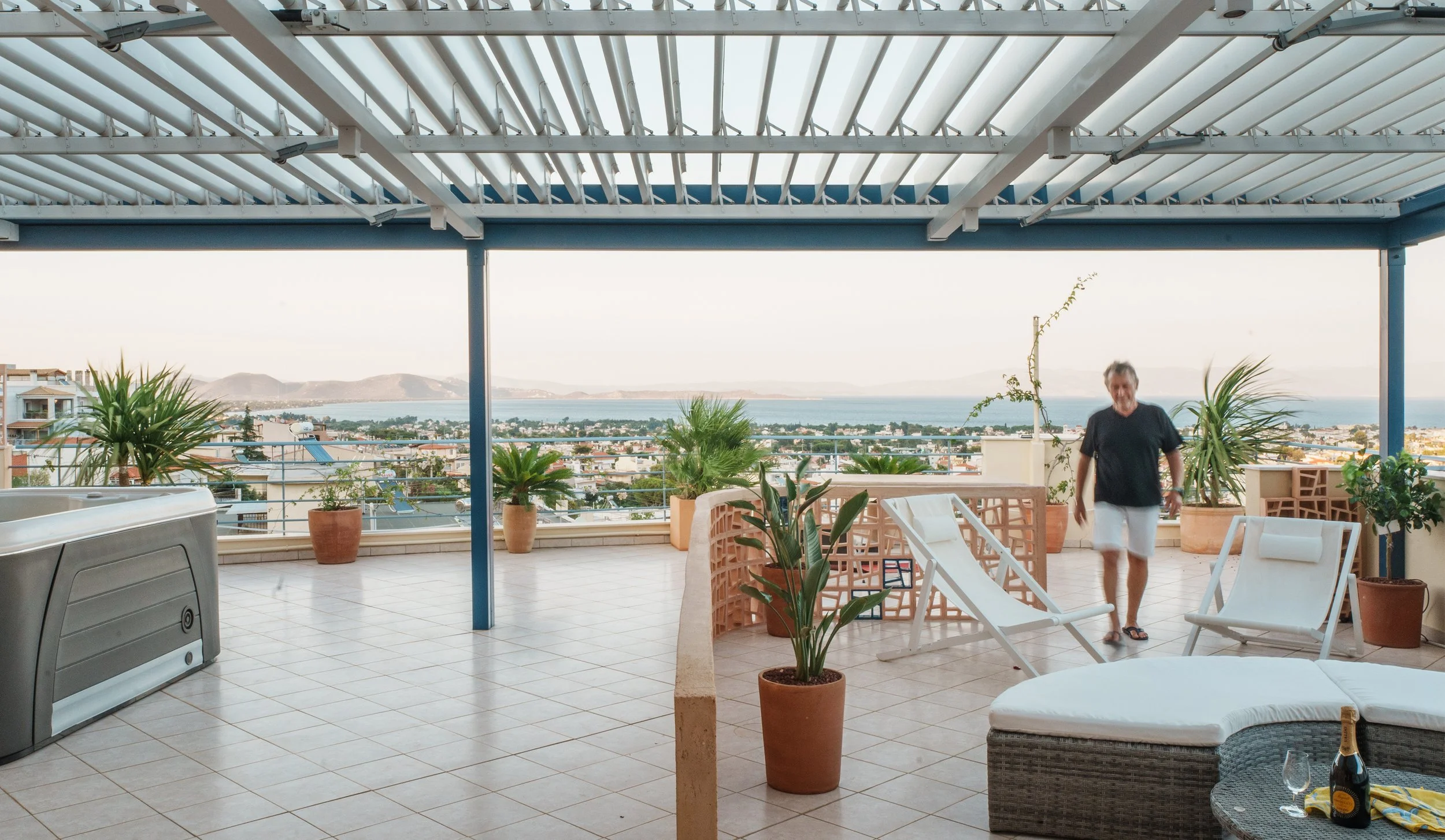 A spacious rooftop patio with potted plants, poolside furniture, and an outdoor grill, overlooking a coastal town with mountains in the distance under a cloudy sky. A man walks across the patio wearing a black shirt, white shorts, and flip-flops.