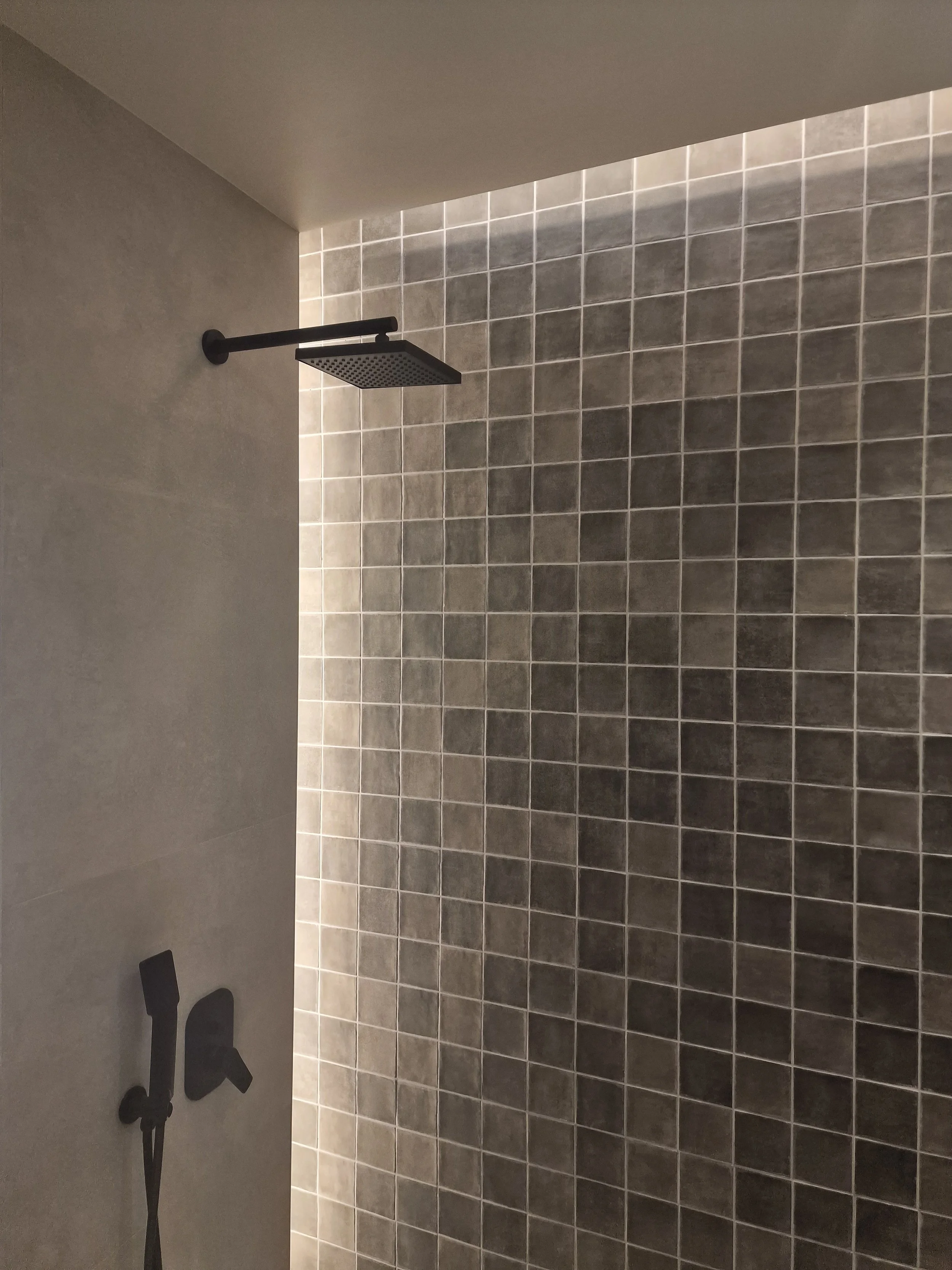 A minimalistic shower area with a square rainfall showerhead mounted on the wall, dark gray tiles on the shower wall, and a partial view of a grey wall with black decorative elements or fixtures.