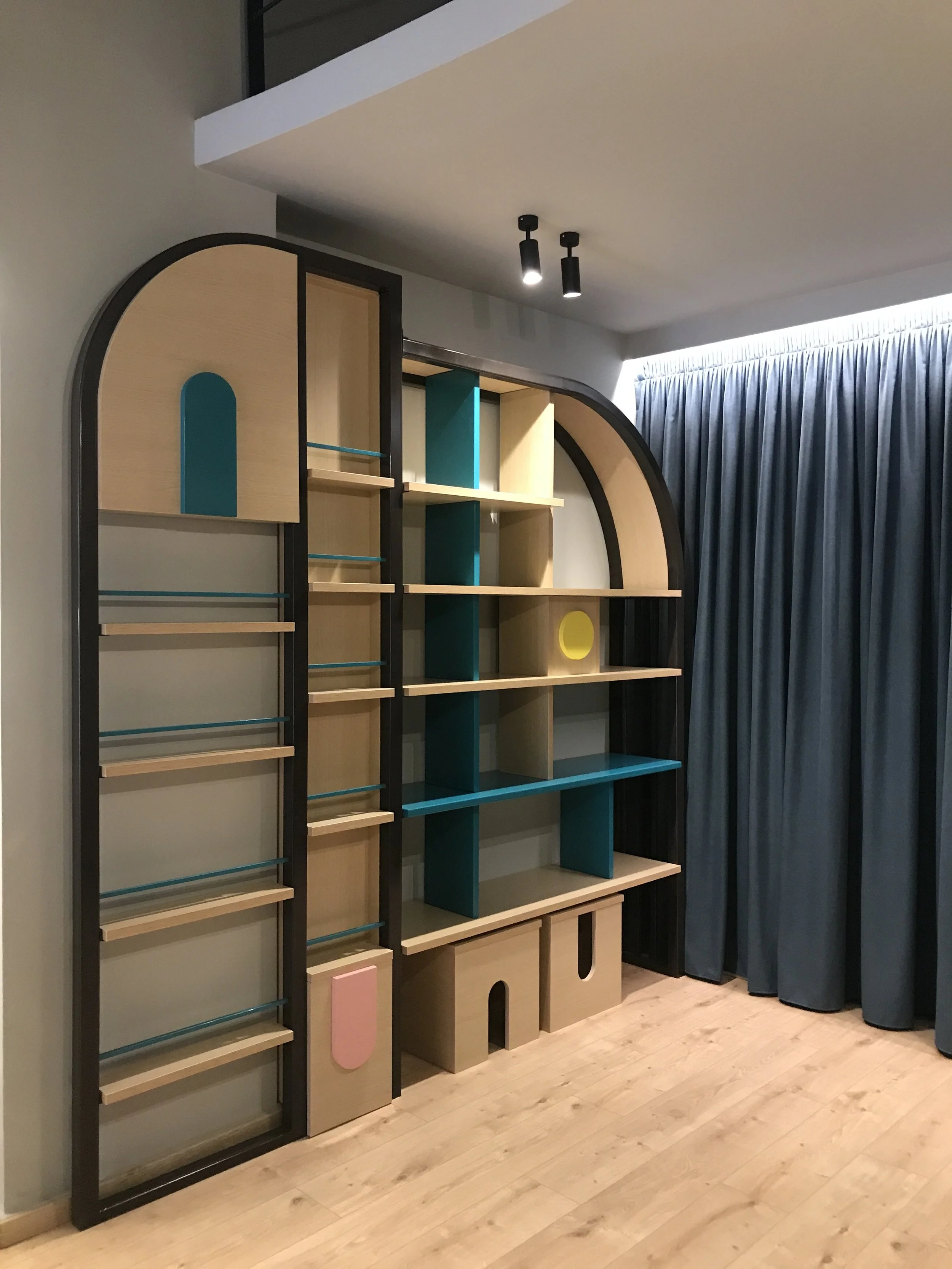 A modern, multi-colored bookshelf with geometric shapes and a combination of open shelves and closed compartments, positioned against a wall with a curtain on the right side and a light wood floor beneath.