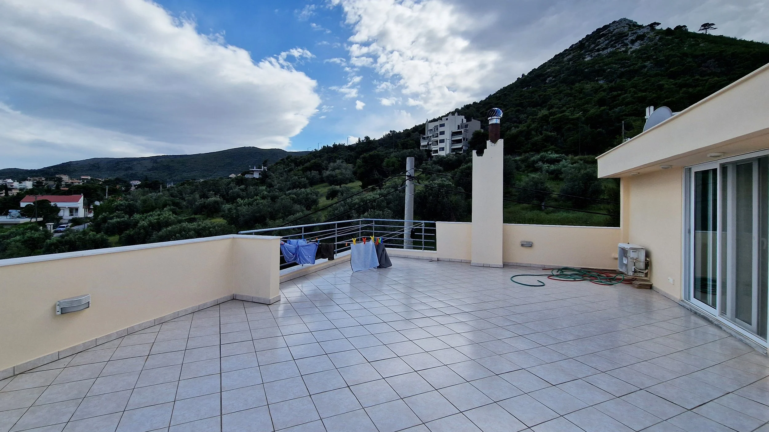 Balcony with tiled floor, laundry hanging to dry, and a mountain view under a cloudy sky, part of a modern apartment building.