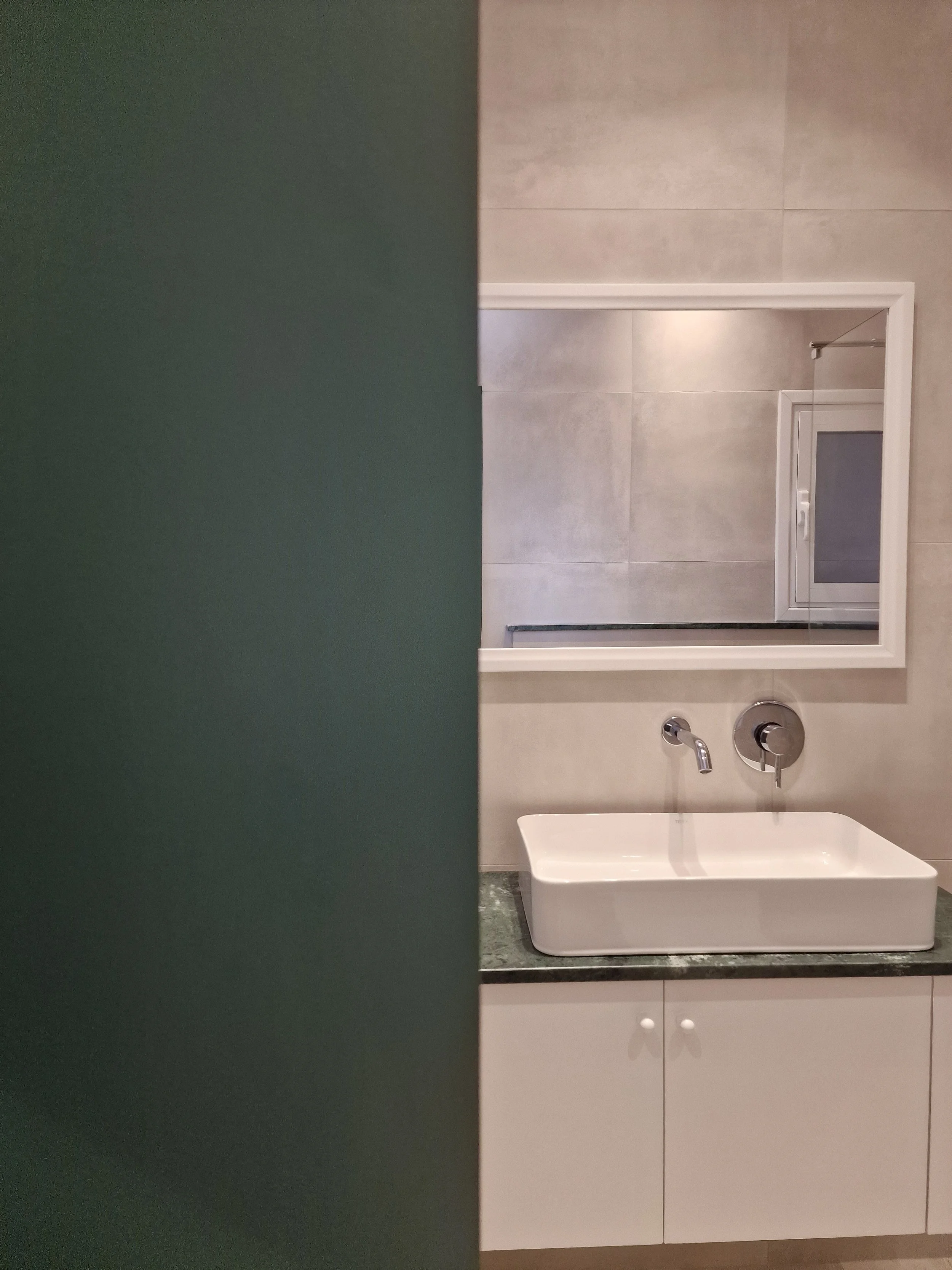 Bathroom with beige tiled wall, white cabinet with white doors, a rectangular white sink, a round wall-mounted faucet, a mirror, and a closed window.