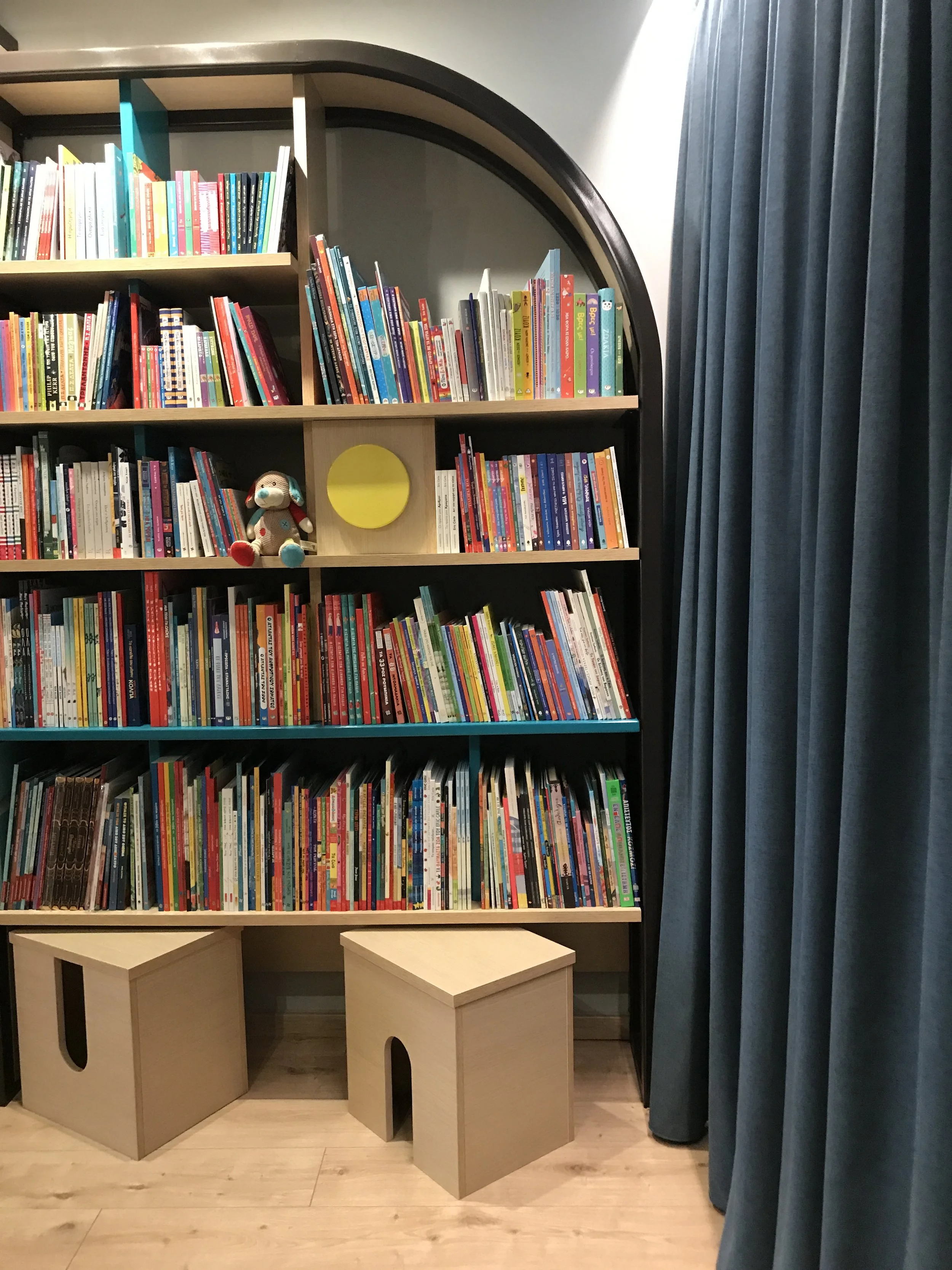 Bookcase filled with children's books, with a stuffed animal and a yellow round object on the middle shelf, and two small wooden stools in front, next to dark blue curtains.