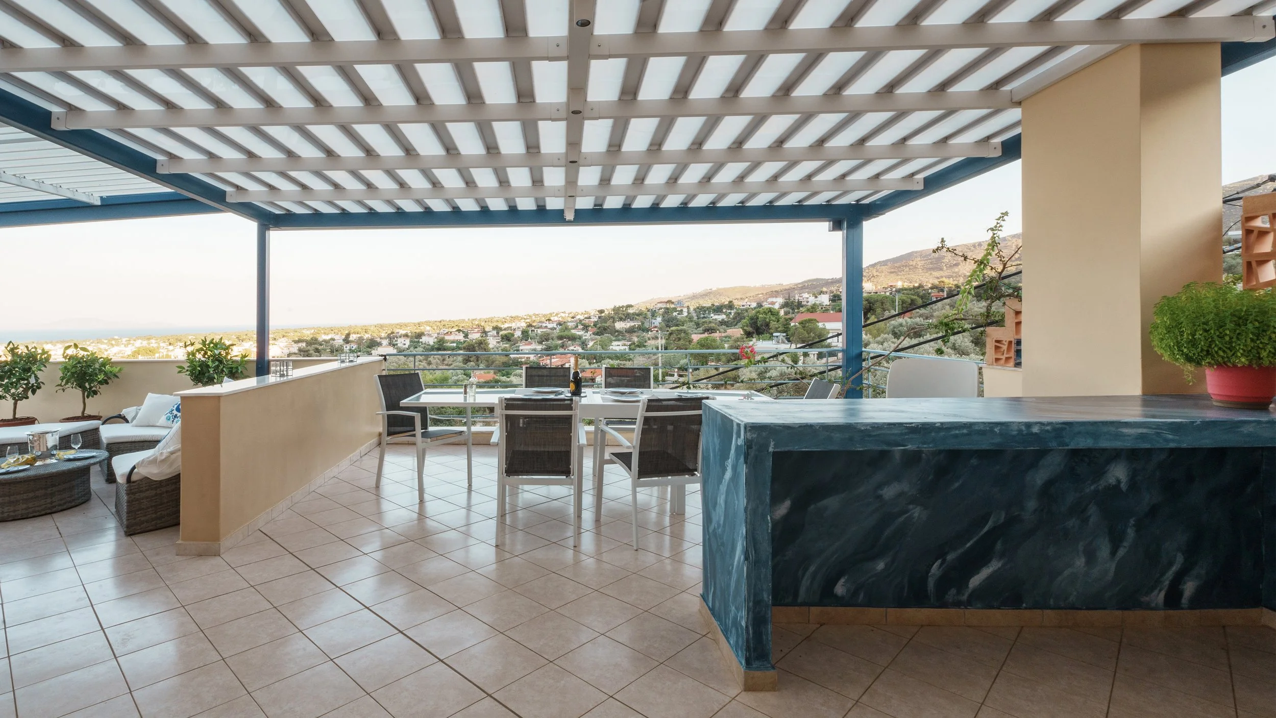 An outdoor patio with a dining table, chairs, a lounge area, and potted plants, overlooking a hilly landscape and sky in the background.