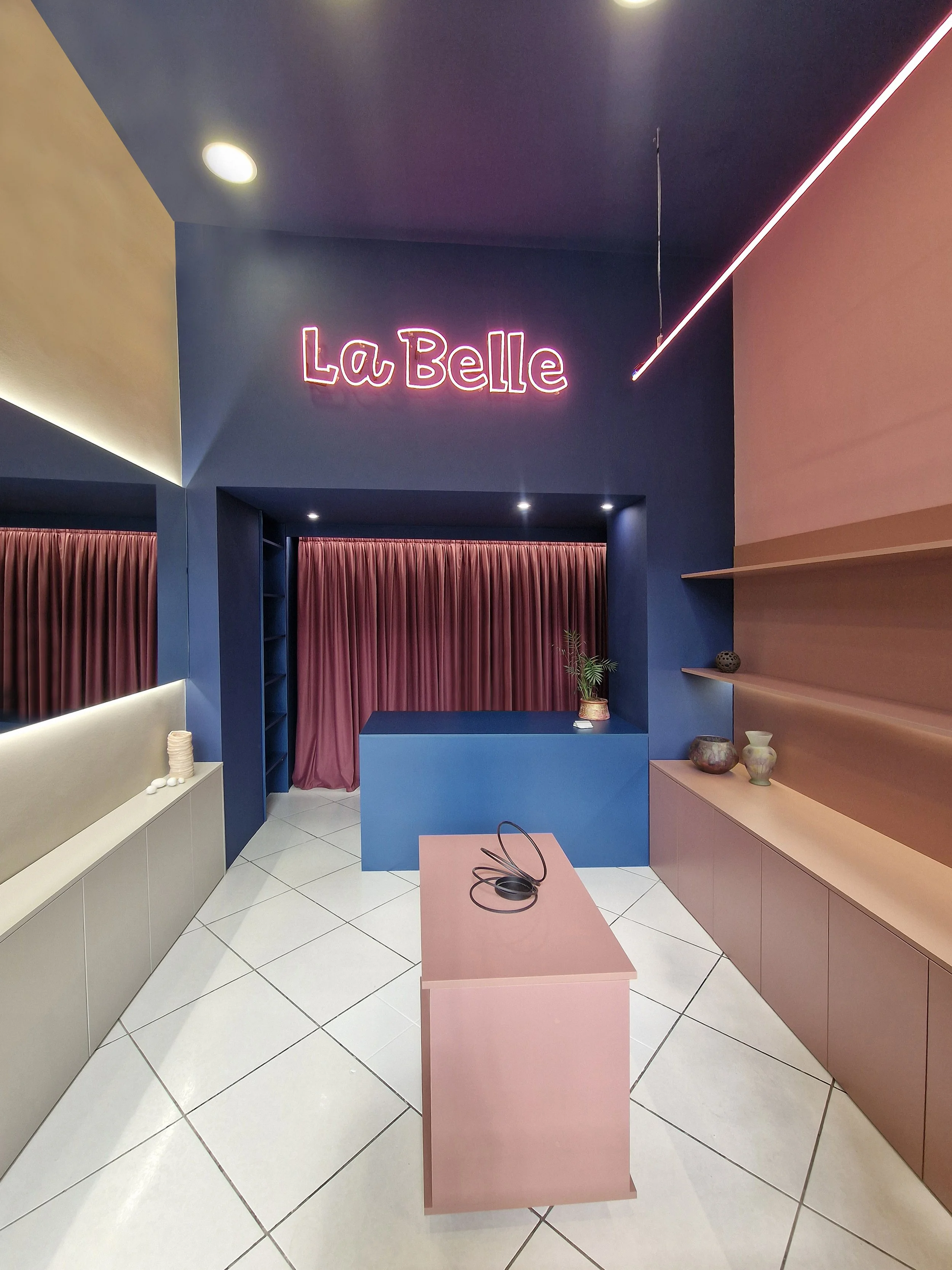 Interior of a modern boutique or store with pink, blue, and beige colored walls, a pink table with a pair of black eyeglasses on it, and decorative pottery on built-in shelves. There's a purple curtain and a neon sign that reads 'La Belle' on a dark 