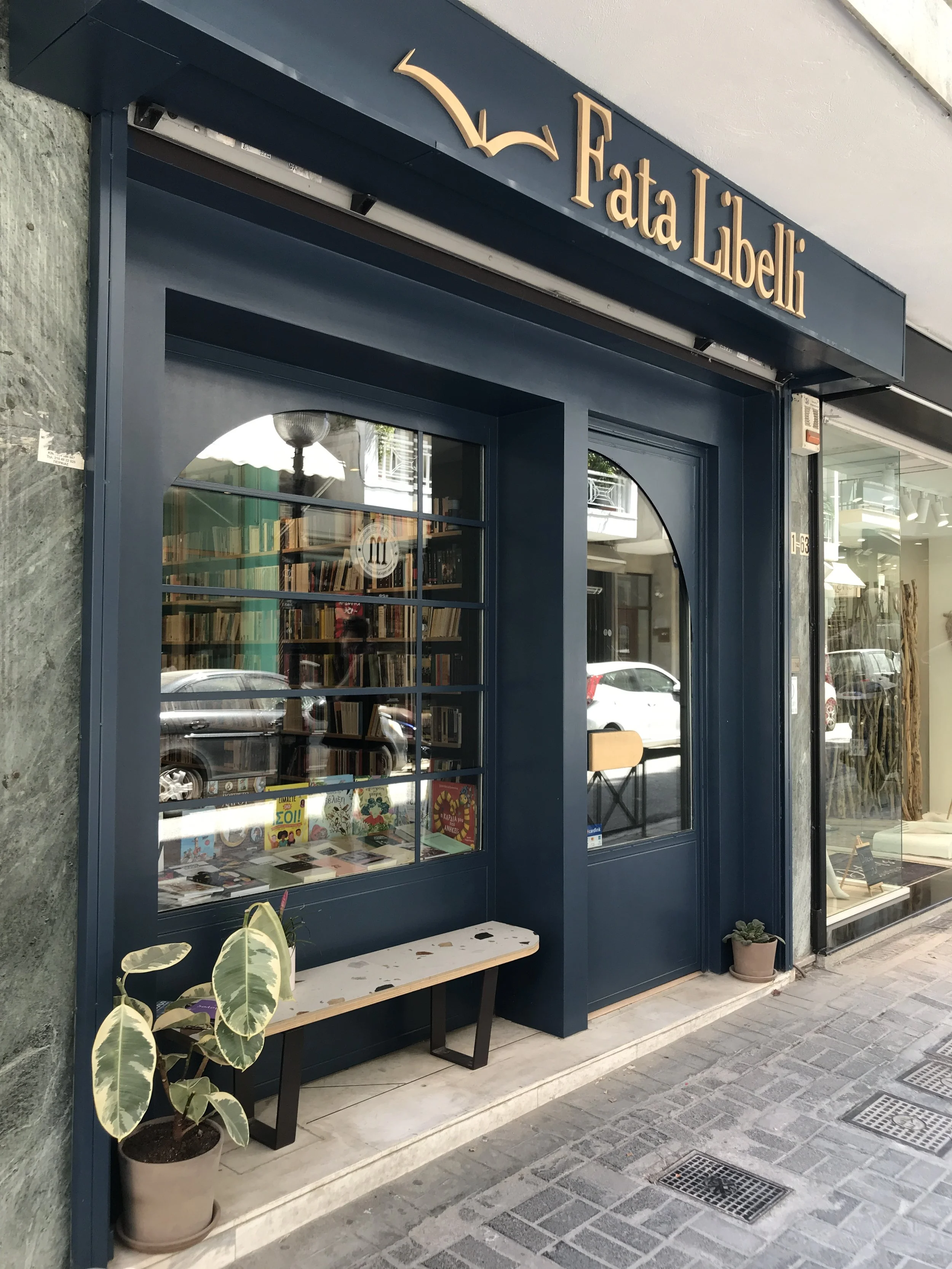 Bookstore entrance with a blue facade, a window display of books and magazines, a small bench, potted plants, and a sign reading 'Data Libelli' with a bird logo above it. The storefront is on a sidewalk with drainage grates.
