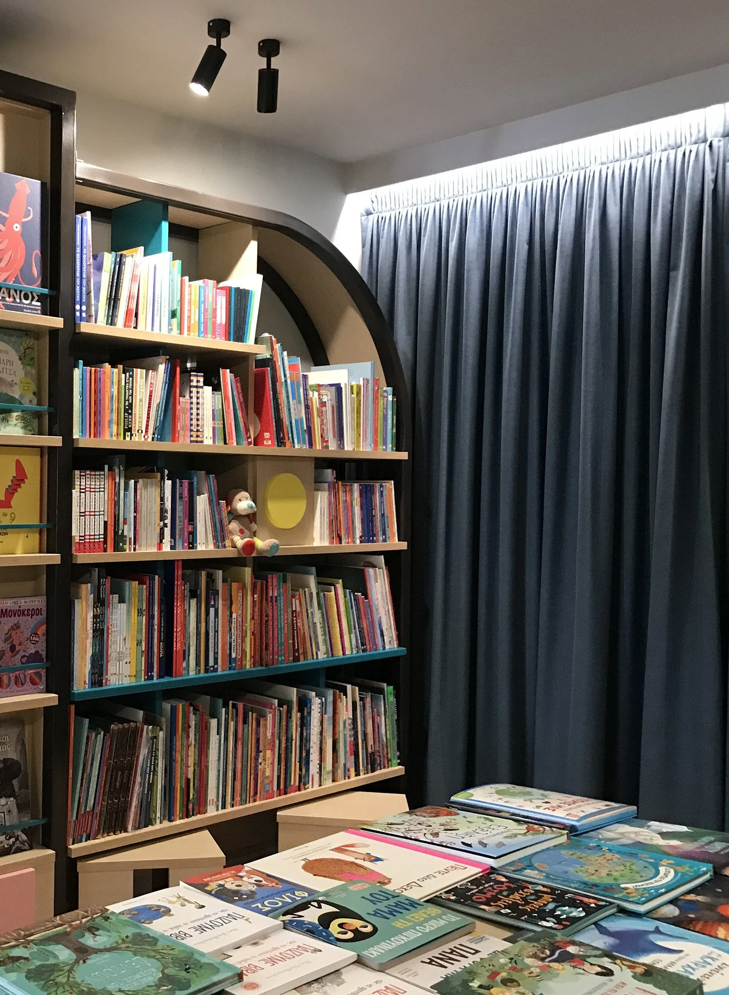 A bookshelf filled with colorful children's books and a table with more children's books on top, with a window covered by dark curtains in the background.