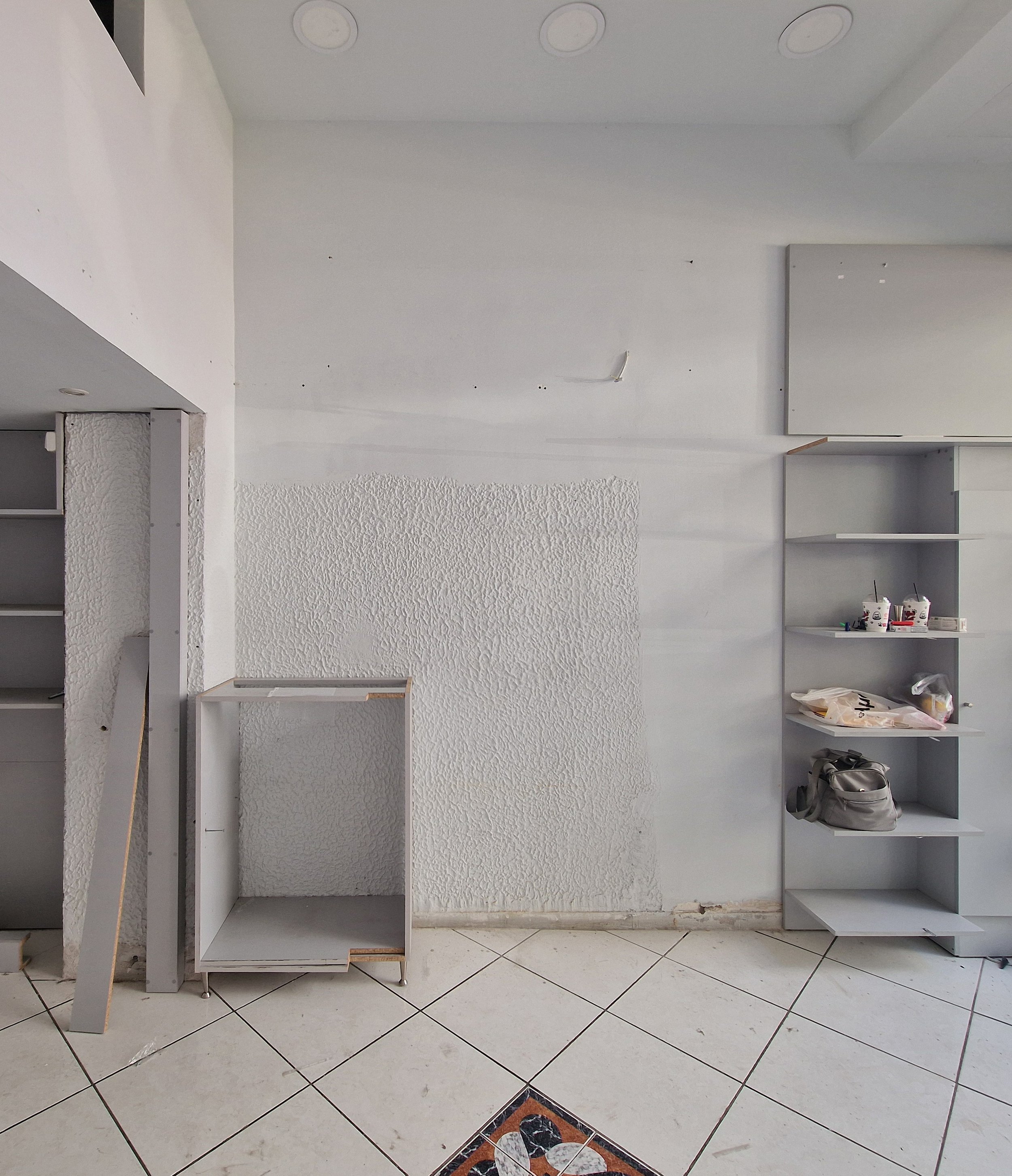 Empty and partially assembled white shelves against a white wall, with some miscellaneous items including cups and bags on the lower shelf, next to a tiled floor with a decorative border.