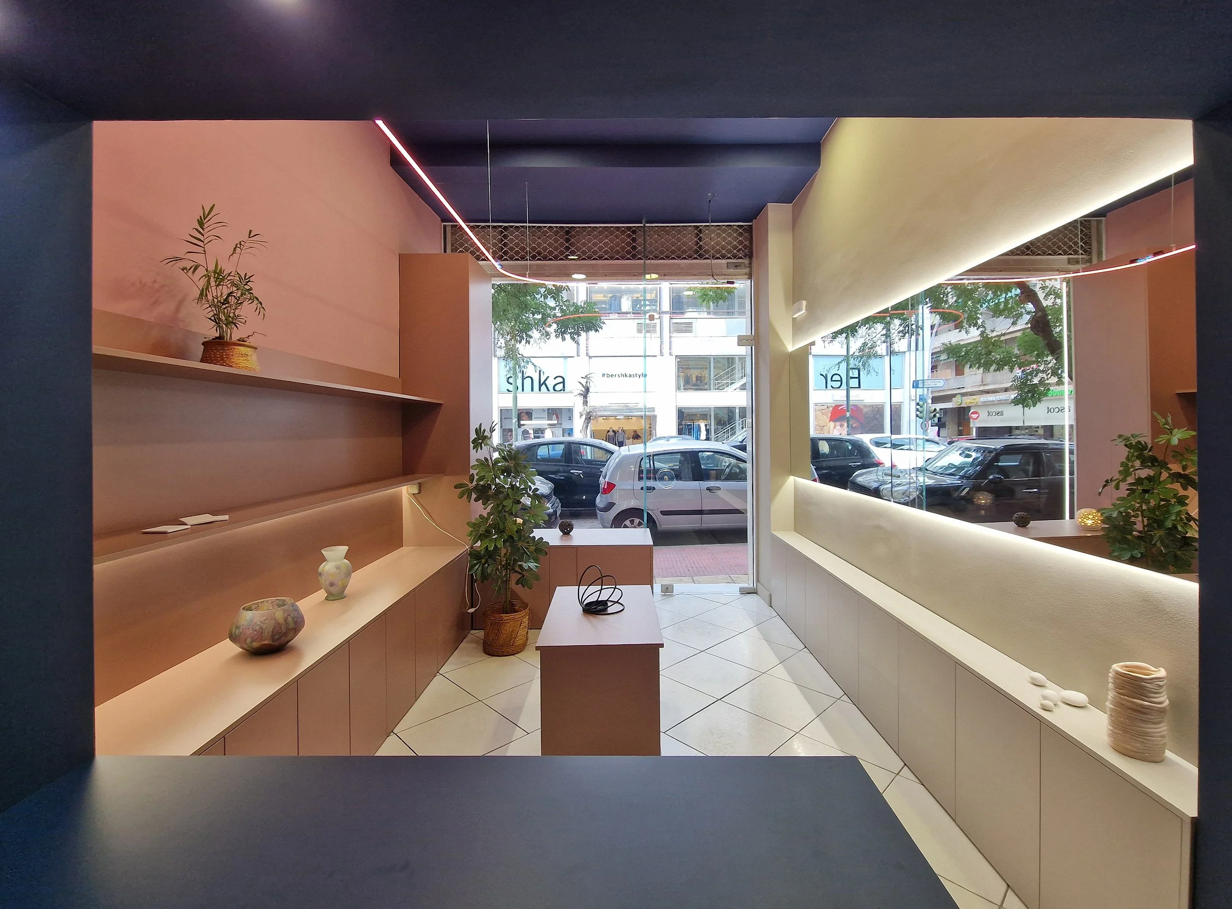 Interior view of a modern space with display shelves holding pottery and plants, large glass window showing parked cars and buildings outside, ceiling with pink neon light accents, and a mix of pink, yellow, and dark blue walls.