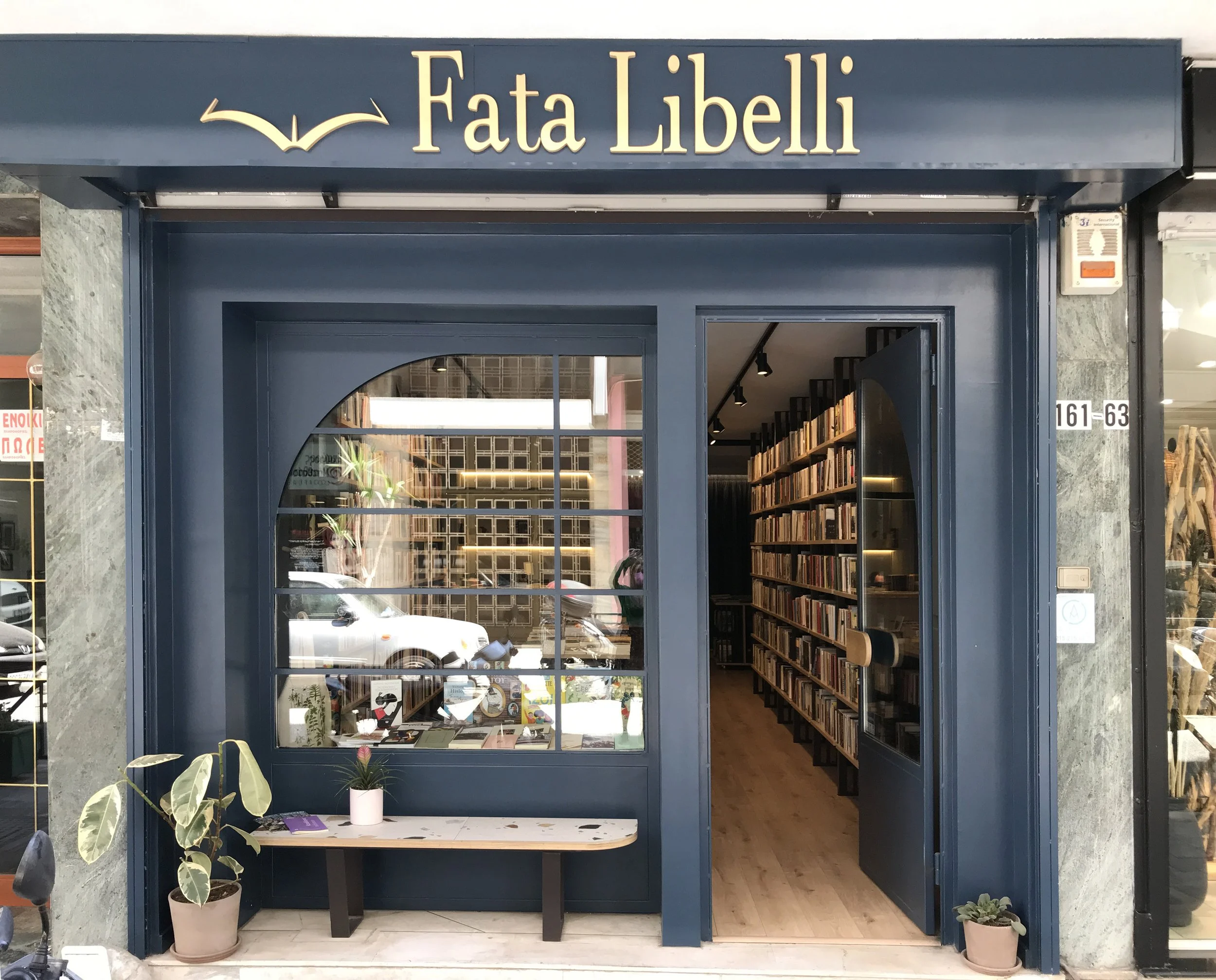 Storefront with blue frame and large window displaying books inside, with potted plants outside and sign reading 'Fata Libelli' with an open book logo.