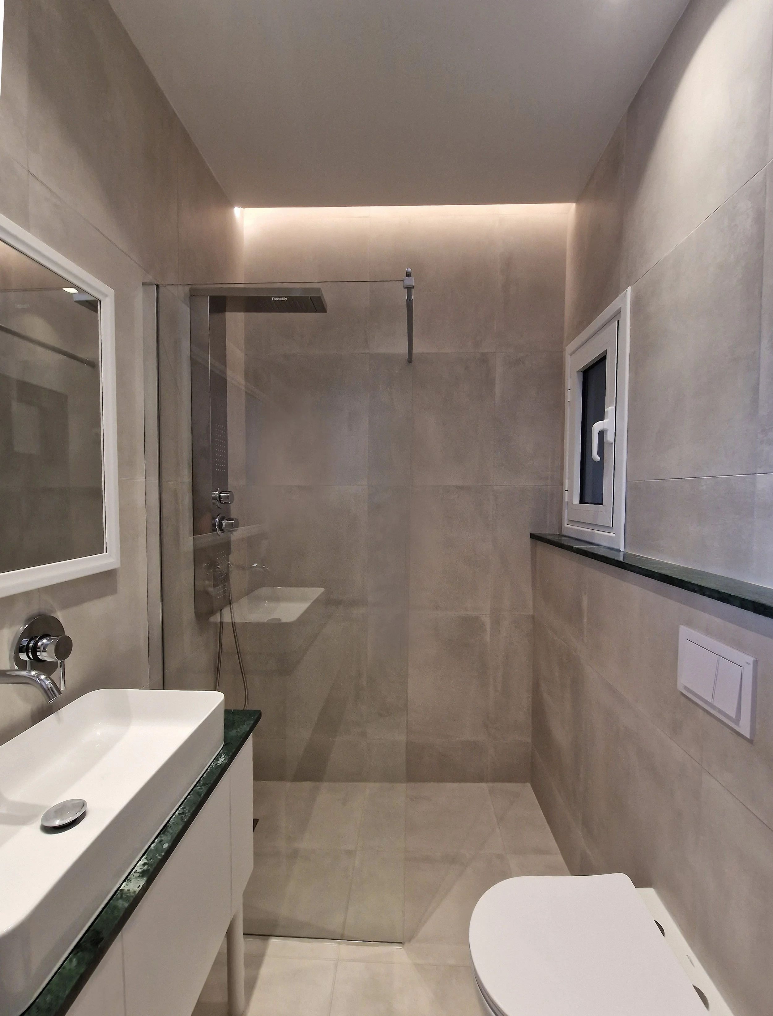 A modern bathroom with a white rectangular sink on a black marble countertop, a wall-mounted faucet, a large mirror, tiled walls, a small window, and a toilet visible in the foreground.