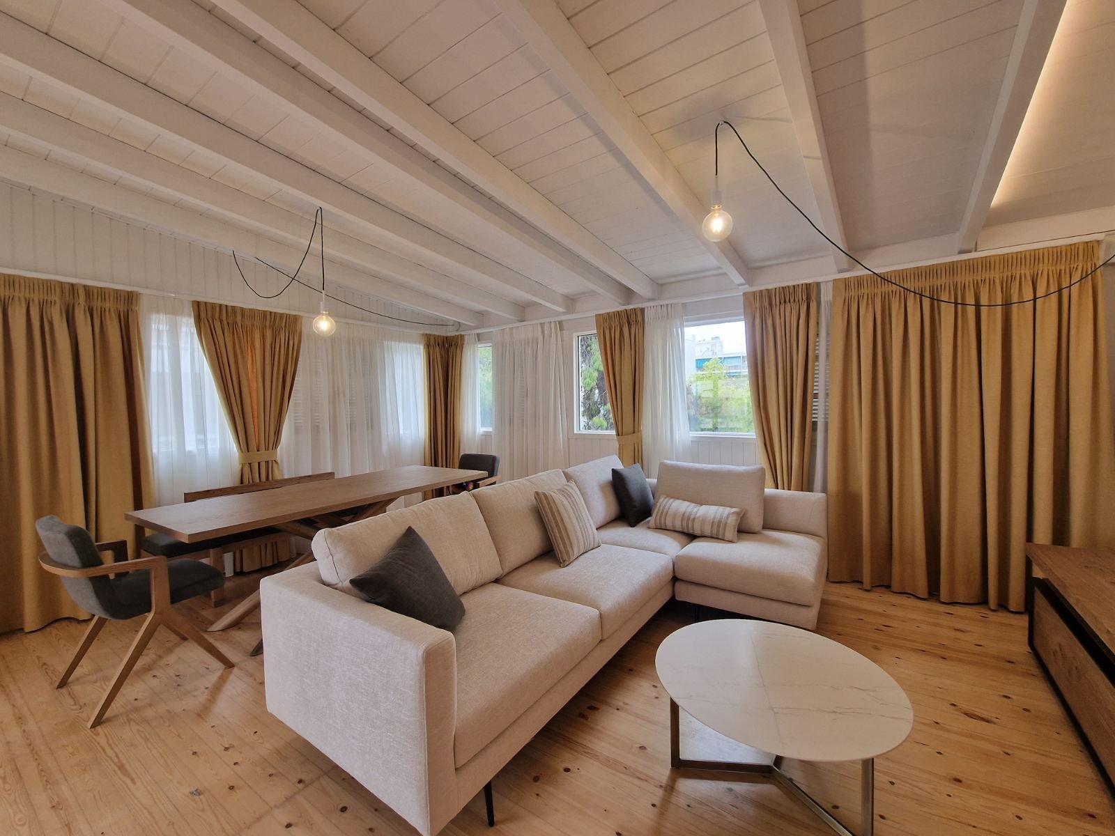 Living room with beige sofa, wooden table, armchairs, multiple windows with white and gold curtains, and wooden floors.