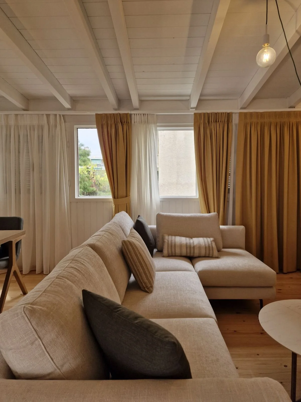 A cozy living room with a beige sectional sofa decorated with striped and solid throw pillows. The room features wooden floors, cream and mustard curtains, and large windows with a view of trees outside. The ceiling has exposed white wooden beams and