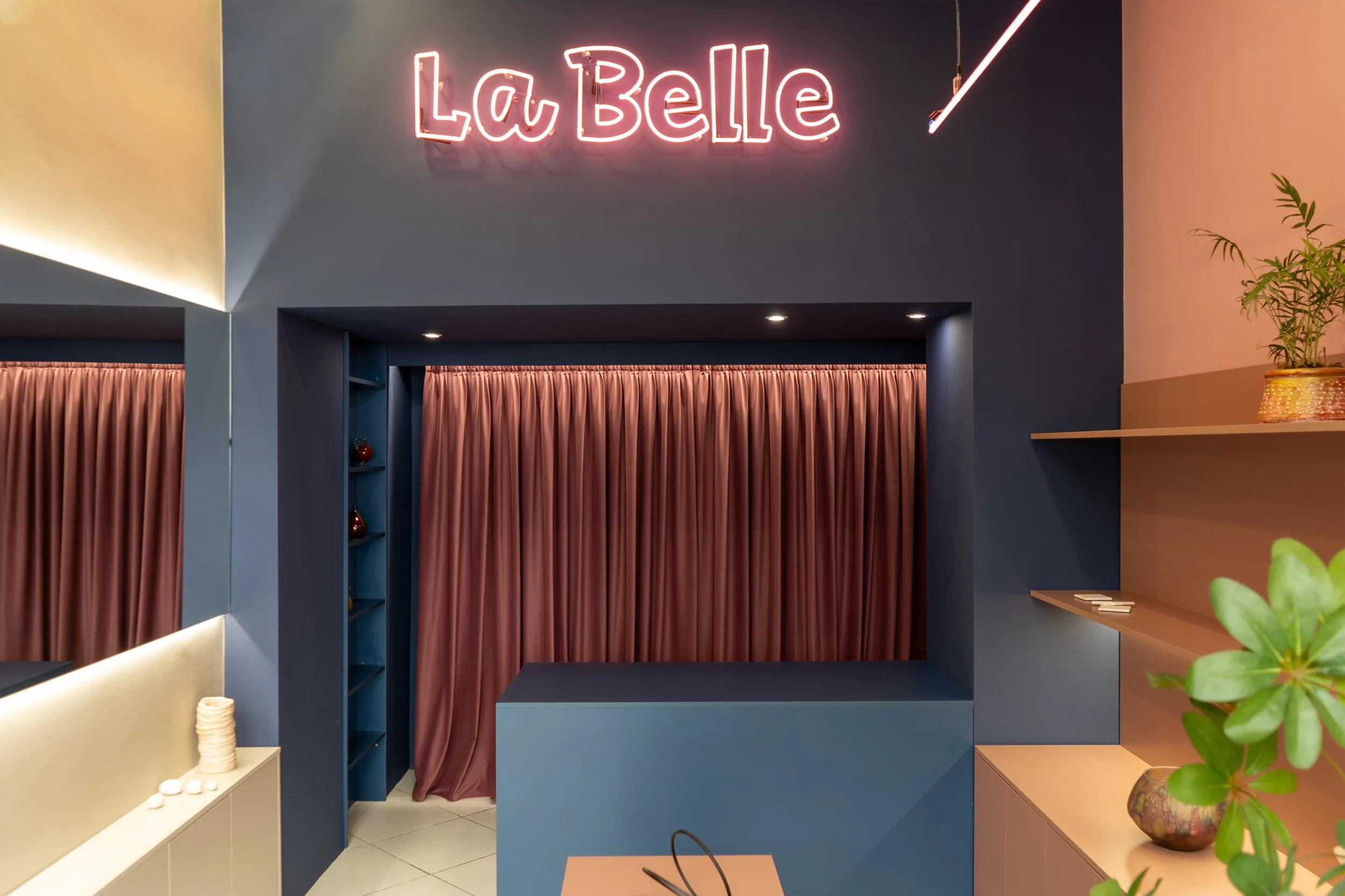 Interior of a stylish space with a neon pink sign saying 'La Belle' on a dark wall, blue and beige counters, pink curtains, and decorative items like a potted plant.