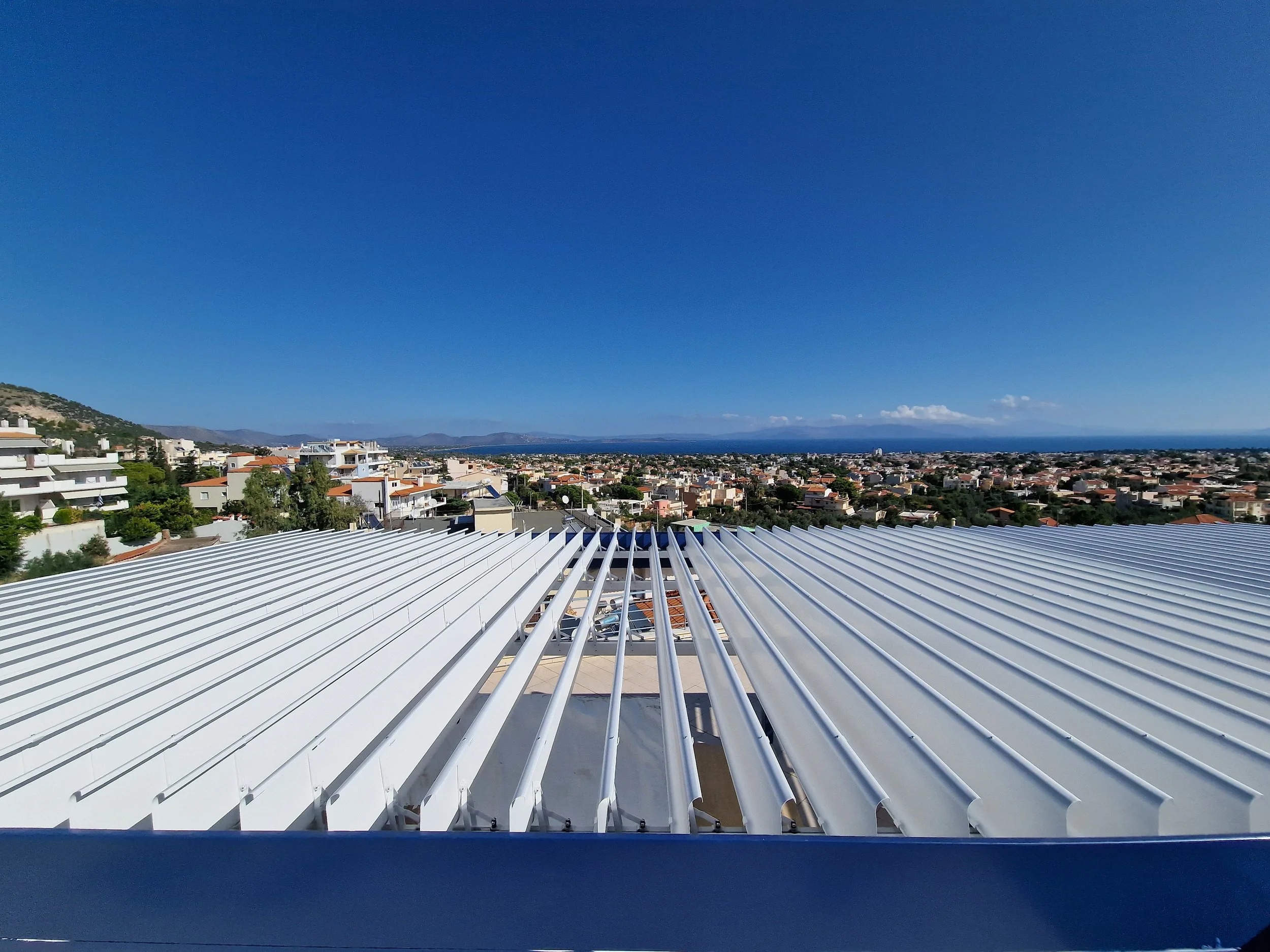 View from a rooftop showing a cityscape with residential buildings, greenery, and distant mountains under a clear blue sky.