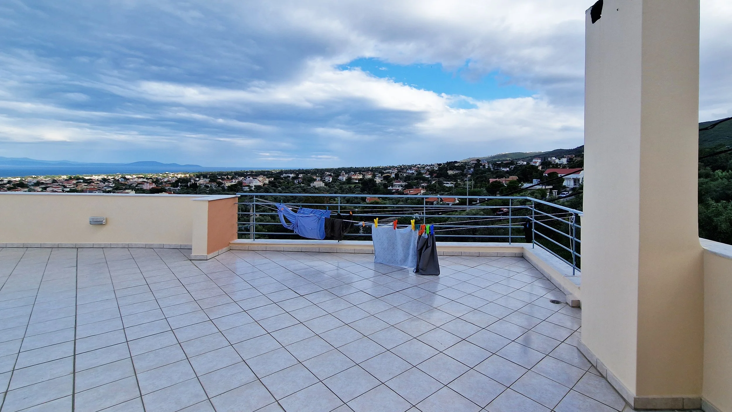 Balcony with laundry hanging on a clothesline overlooking a cityscape, hills, and sky with clouds.