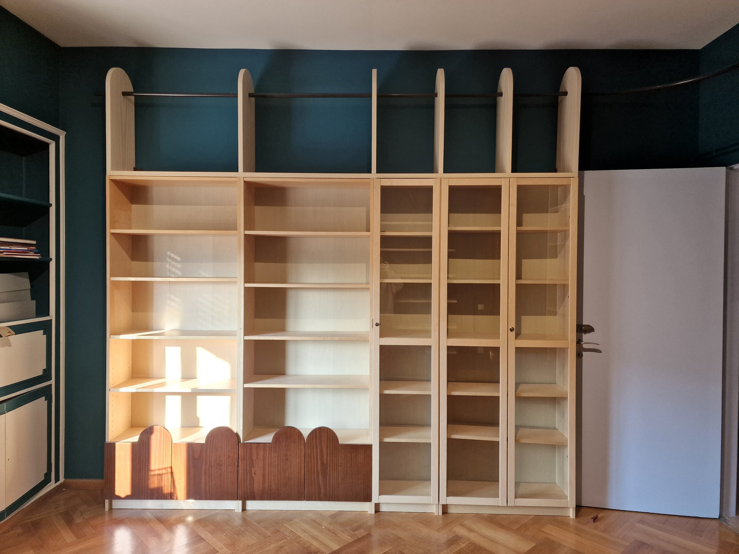 Empty wooden shelving unit with multiple compartments in a room with dark teal wall and hardwood floor.