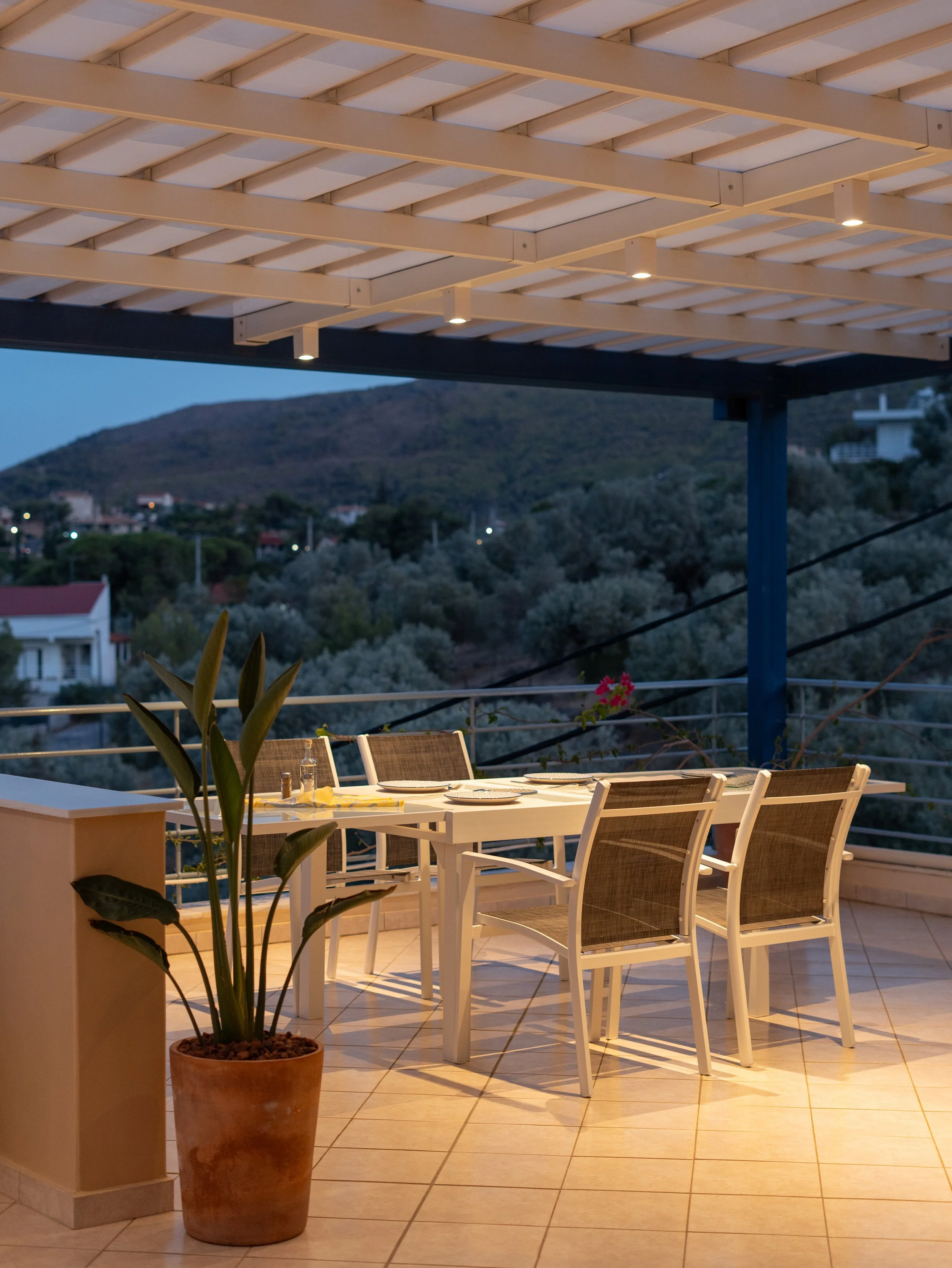 Outdoor dining area with a white table and chairs, potted plant in the foreground, and a scenic view of hills and houses in the evening.