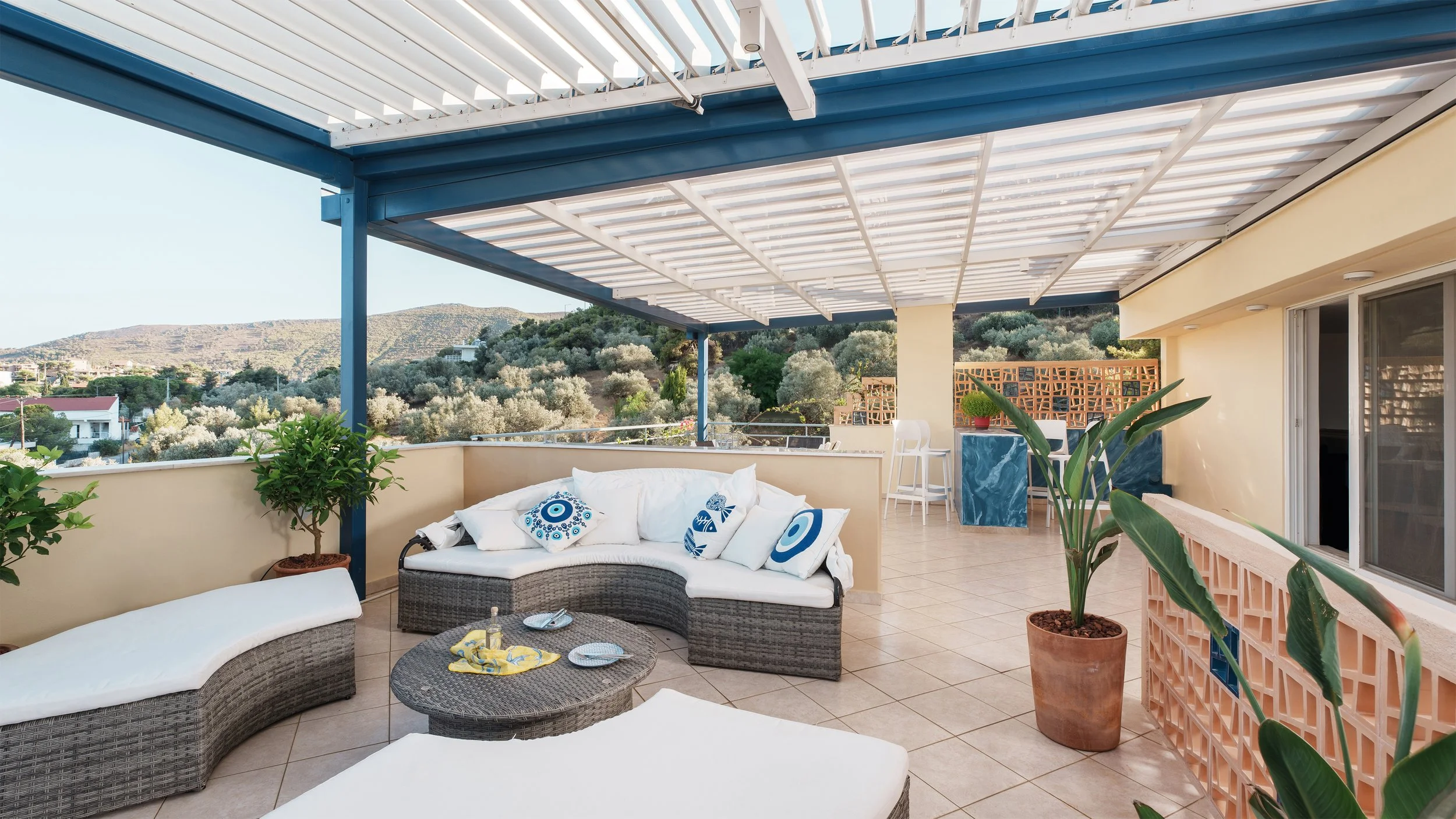 Outdoor rooftop patio with wicker sofa and pillows, potted plants, a small table with dishes, and a view of hills in the background.