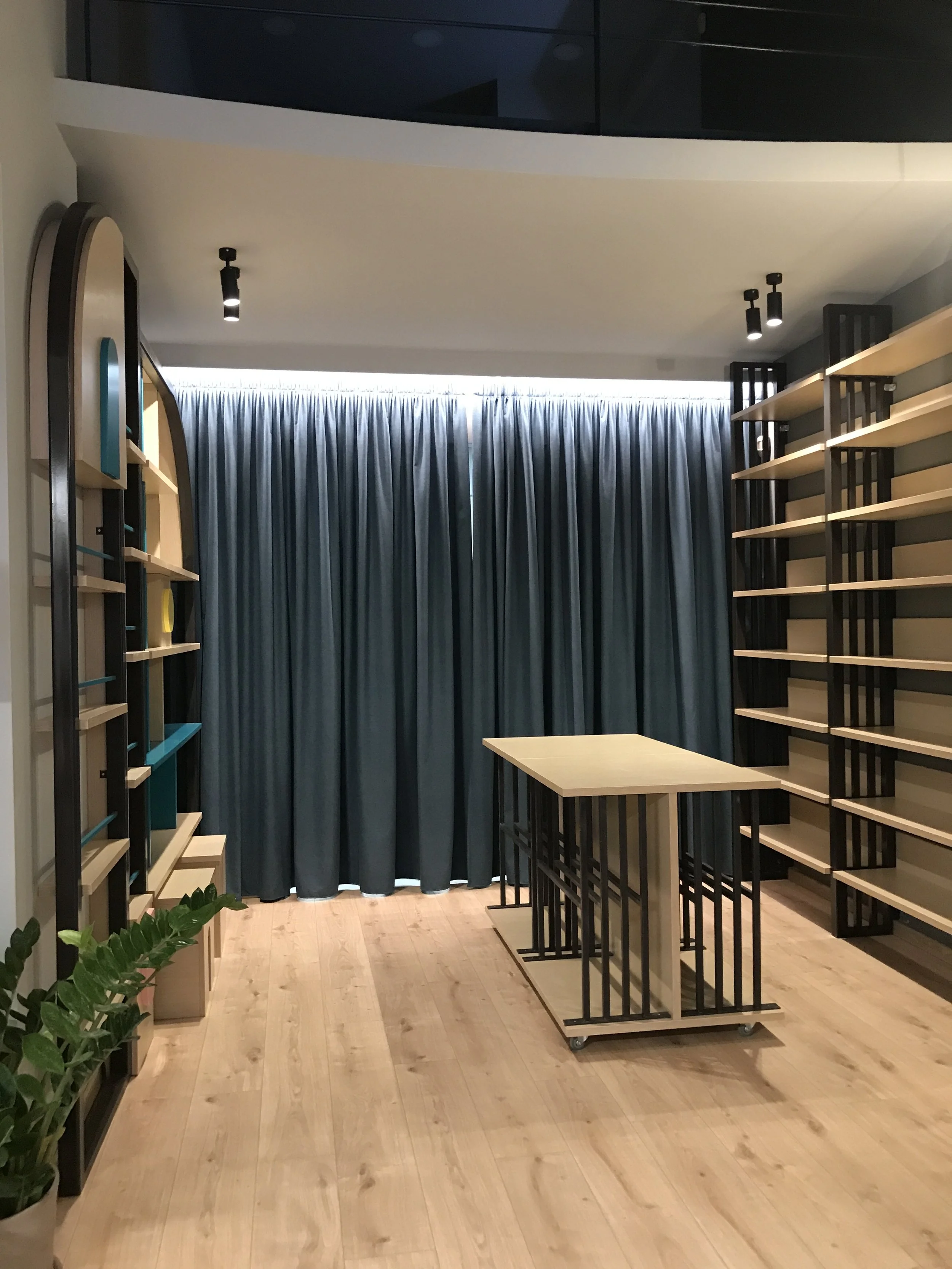 Empty room with wooden floors, tall black metal and light wood shelving units, a small table on wheels, dark blue curtains covering the window, ceiling spotlights, and some green plants in a beige plant holder.