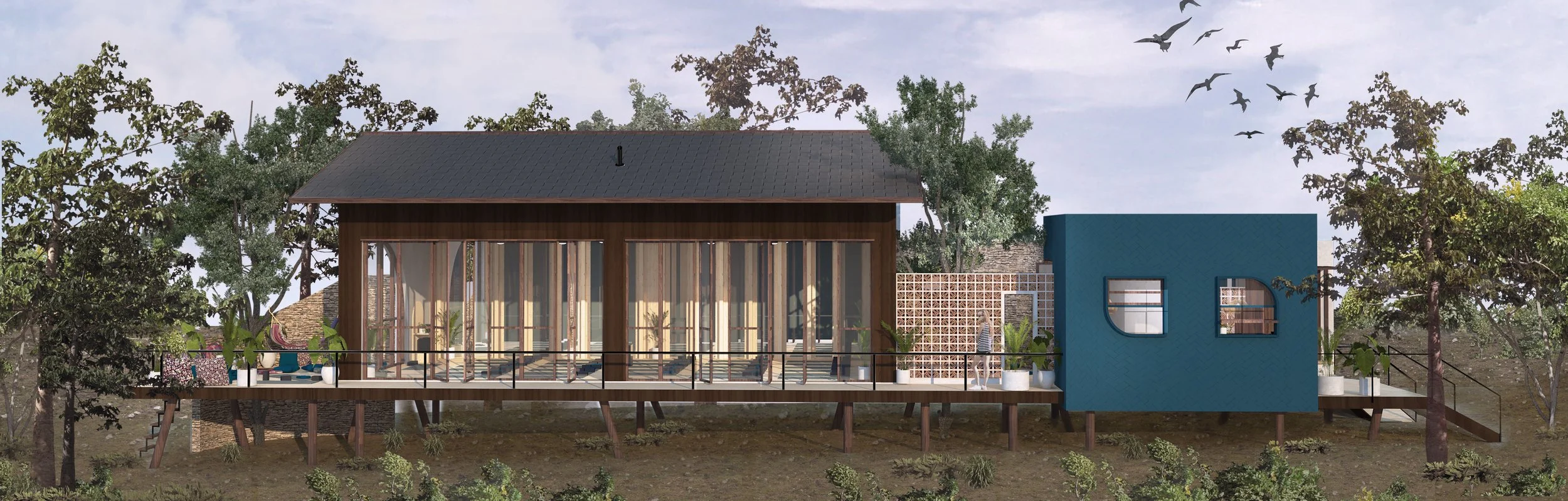 A modern house on stilts with a large glass front, a wooden deck with outdoor furniture, and a small blue addition with windows, surrounded by trees and a cloudy sky.