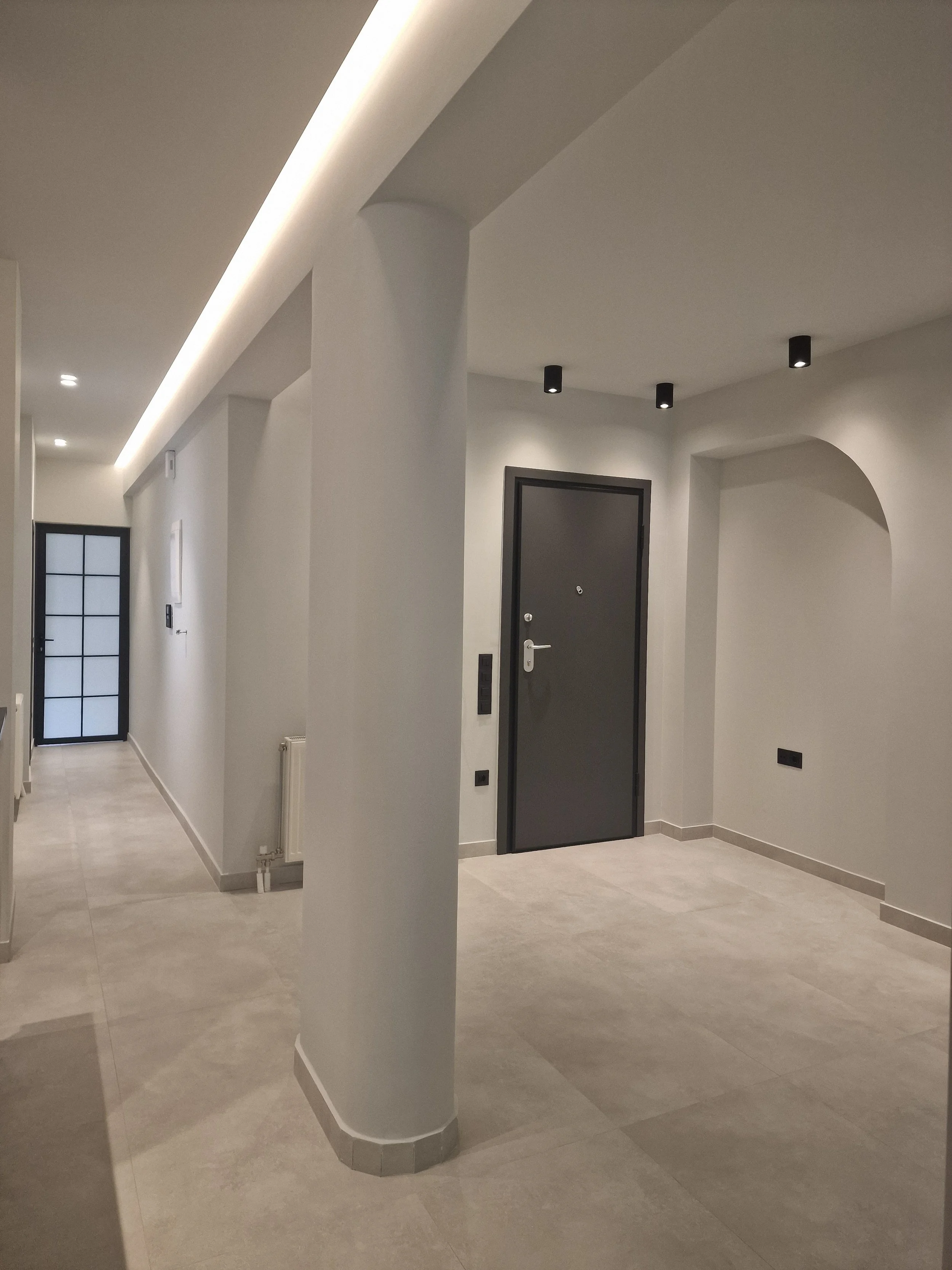 An interior view of a modern, minimalistic hallway with a black door, a window with grid panes, and neutral-toned flooring and walls. The ceiling has recessed and surface-mounted lights.