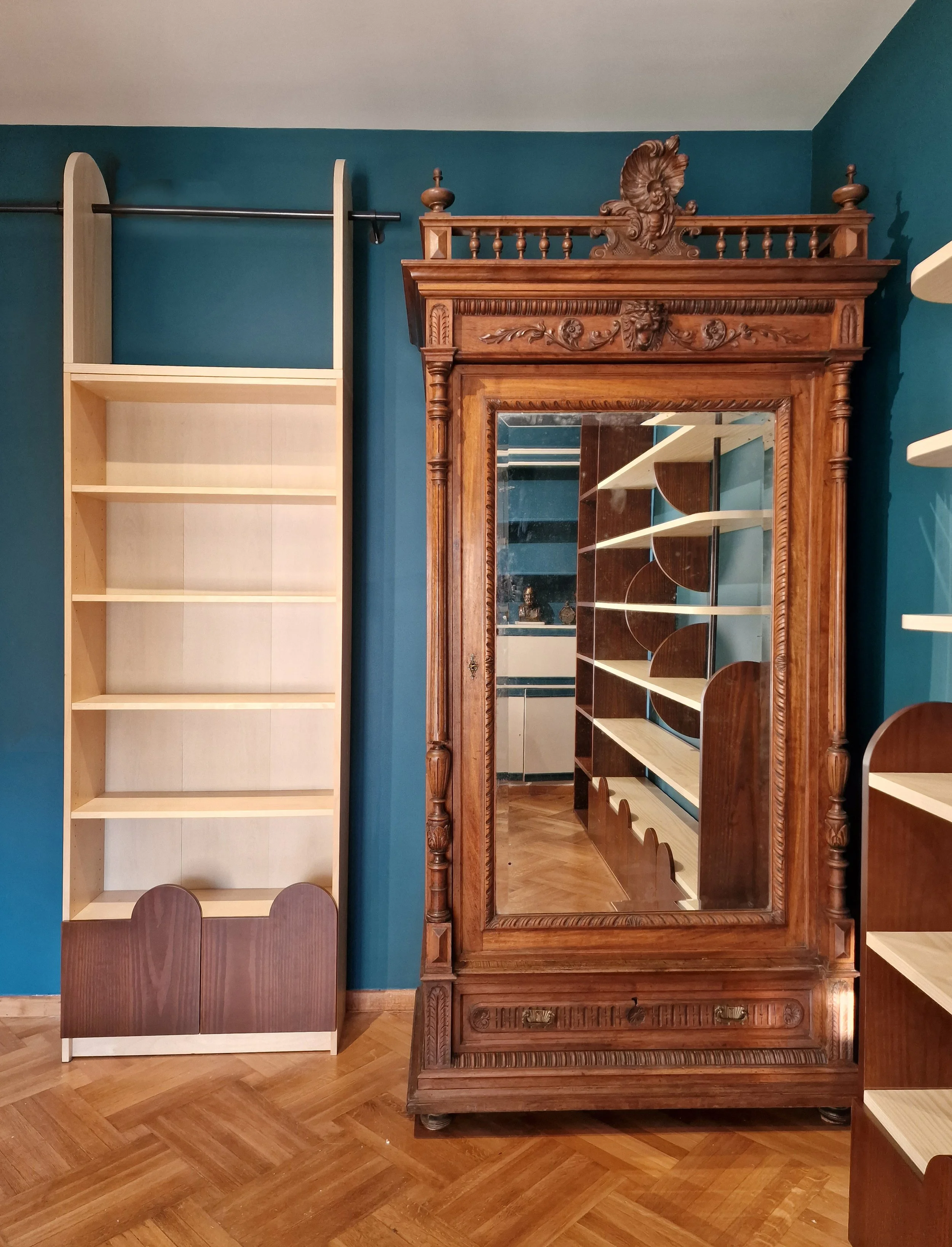 A room with a large ornate wooden mirror with intricate carvings, a wooden cabinet with open shelves, and a plain white and purple bookshelf against a teal wall, with a parquet wood floor.