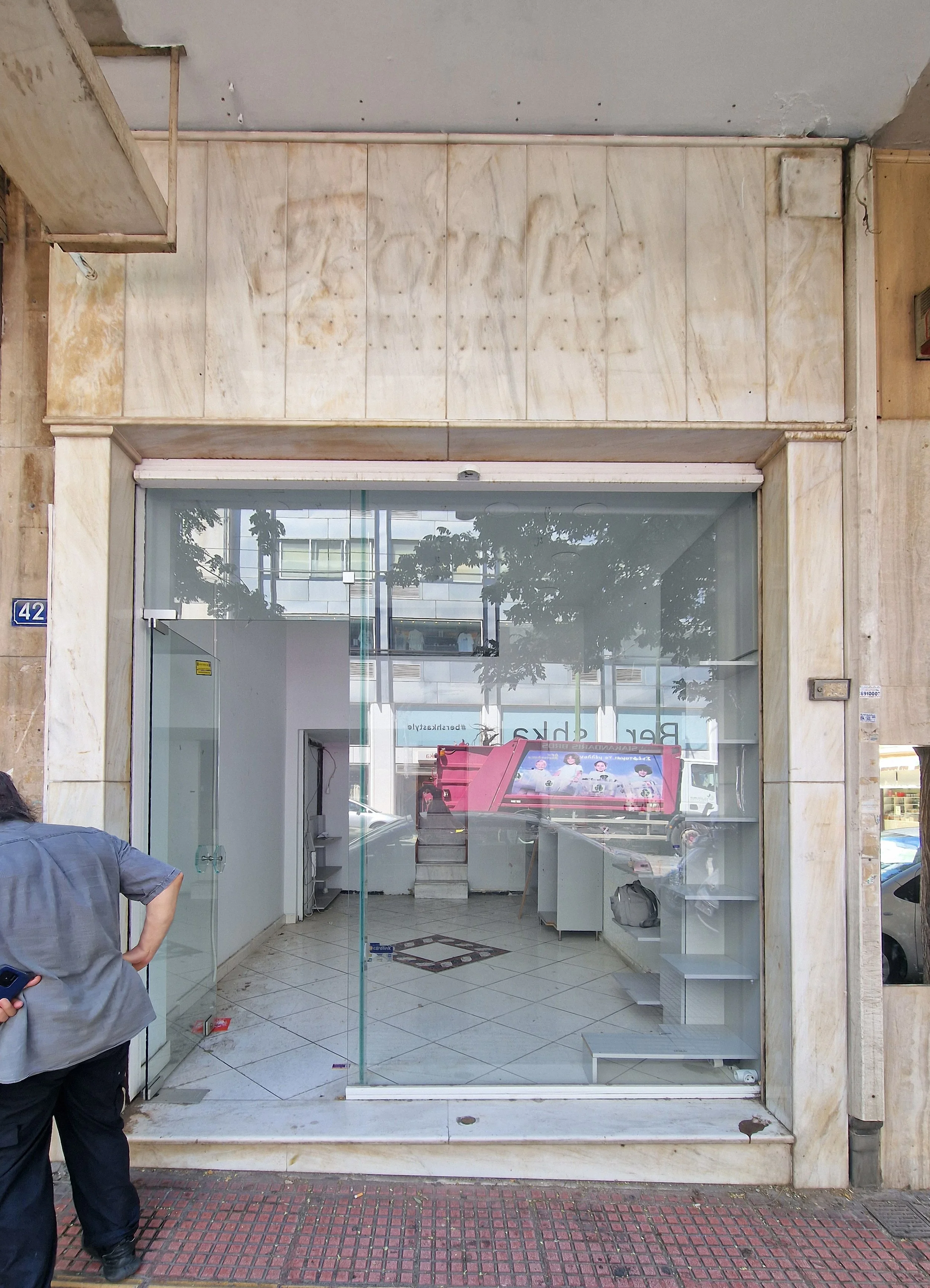 Empty storefront with glass window and door, and a man in gray shirt standing outside. The interior is empty with shelves and a counter, and a pink display stand. The sidewalk is tiled with red and black bricks.