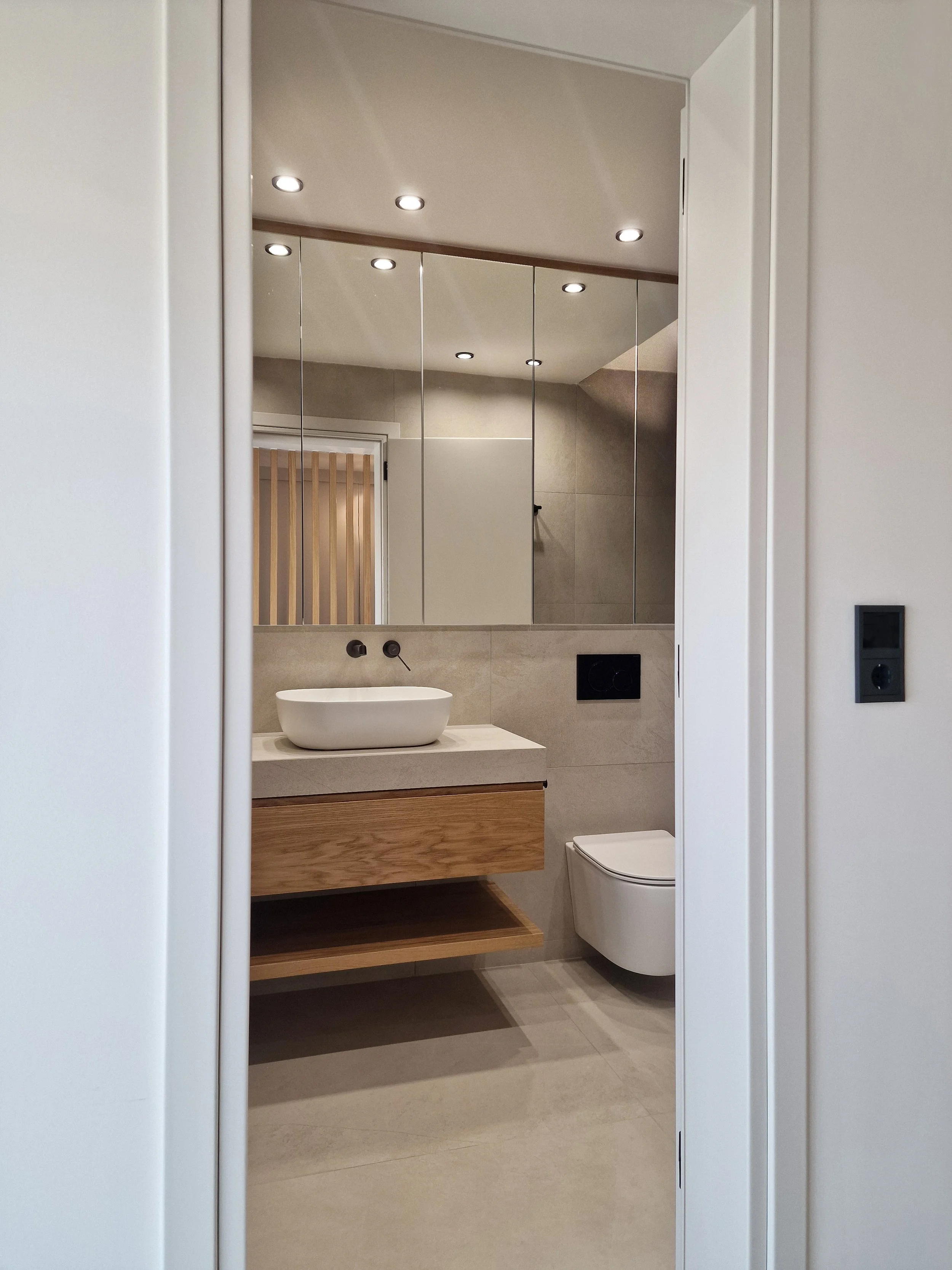 Modern bathroom with a white vessel sink on a wooden vanity, a wall-mounted toilet, large mirror panels, and recessed ceiling lights.