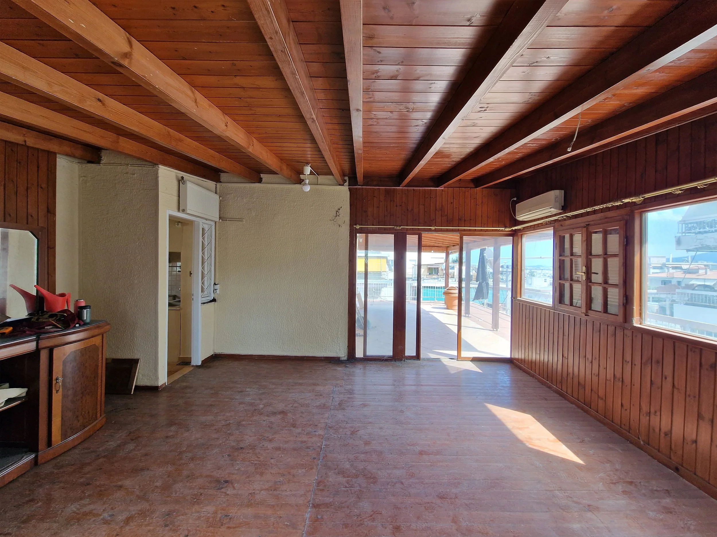 A room with wooden ceiling beams and wood-paneled walls, bright sunlight coming through glass doors leading to an outdoor balcony with a potted plant.