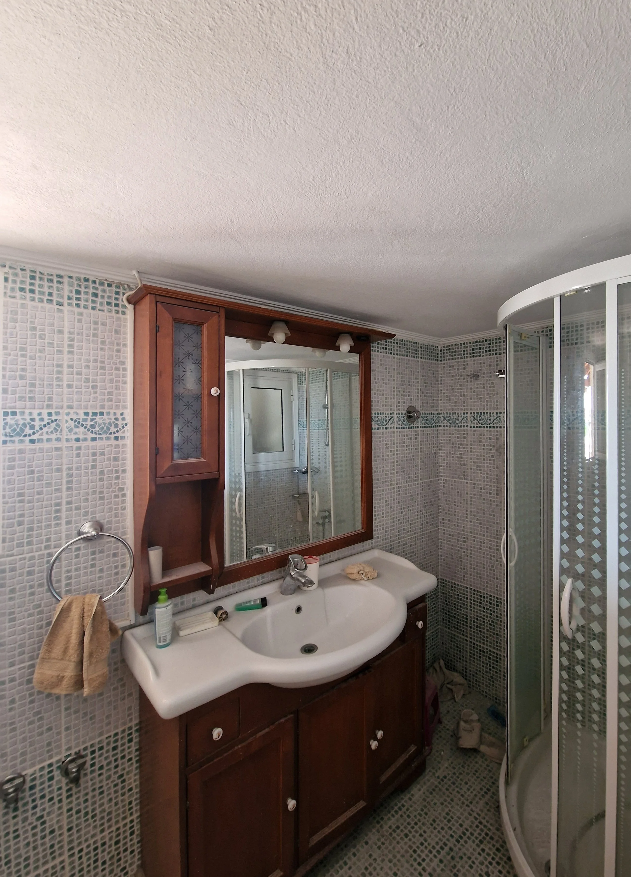 A small bathroom with a white sink, wooden cabinet mirror, mosaic tile walls, and a curved glass shower enclosure.