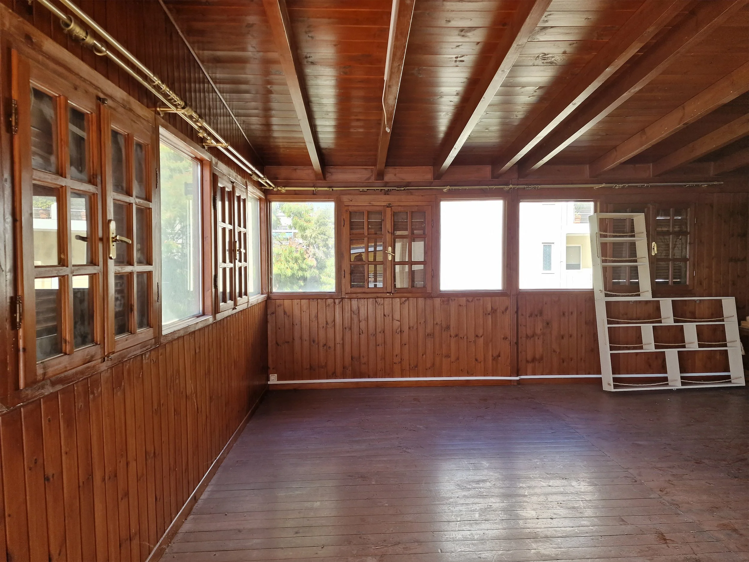 An empty room with wooden walls, ceiling, and floor, featuring large windows with multiple panes and a ladder.