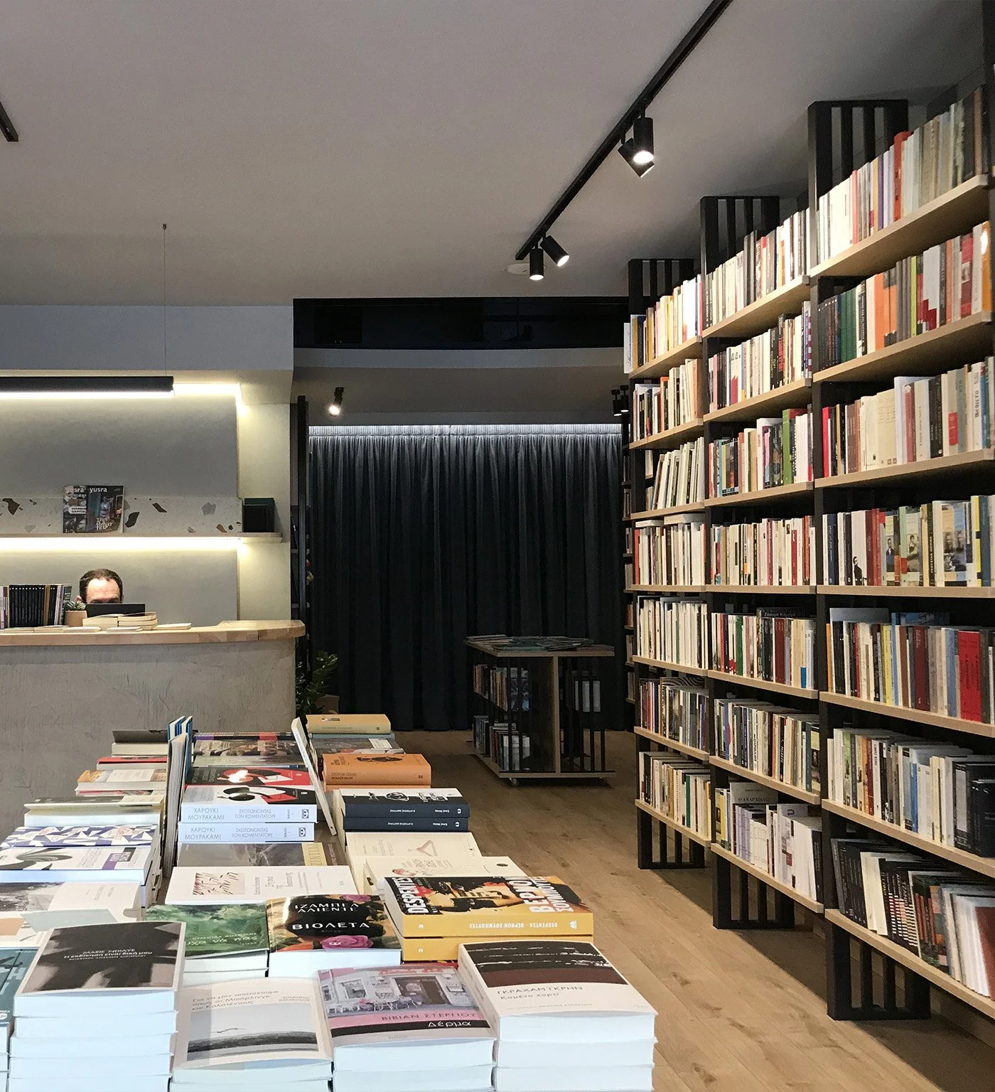 Interior of a bookstore or library with shelves filled with books, a table with books in the foreground, and a checkout counter with a person behind it.