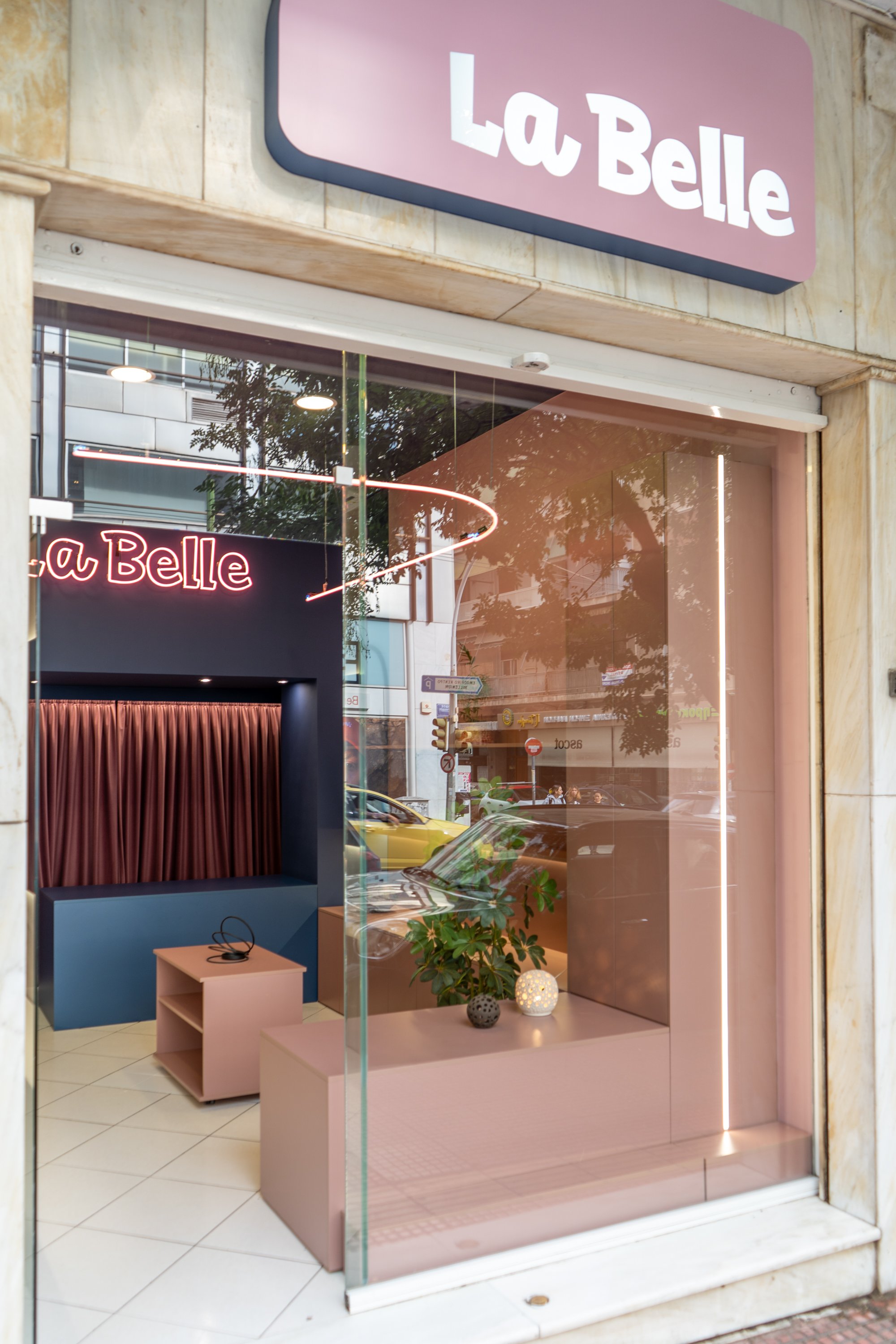Storefront of La Belle with pink and purple sign, large glass window, interior display with pink and purple decor, a small table and decorative objects, plant near window, street view reflections.