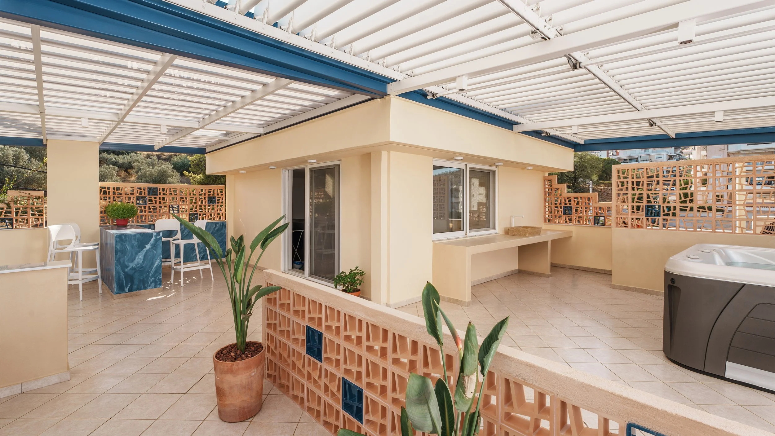 Indoor terrace with beige walls, potted plants, white chairs, a blue marble table, sliding glass doors, and a rooftop pergola with white slats.