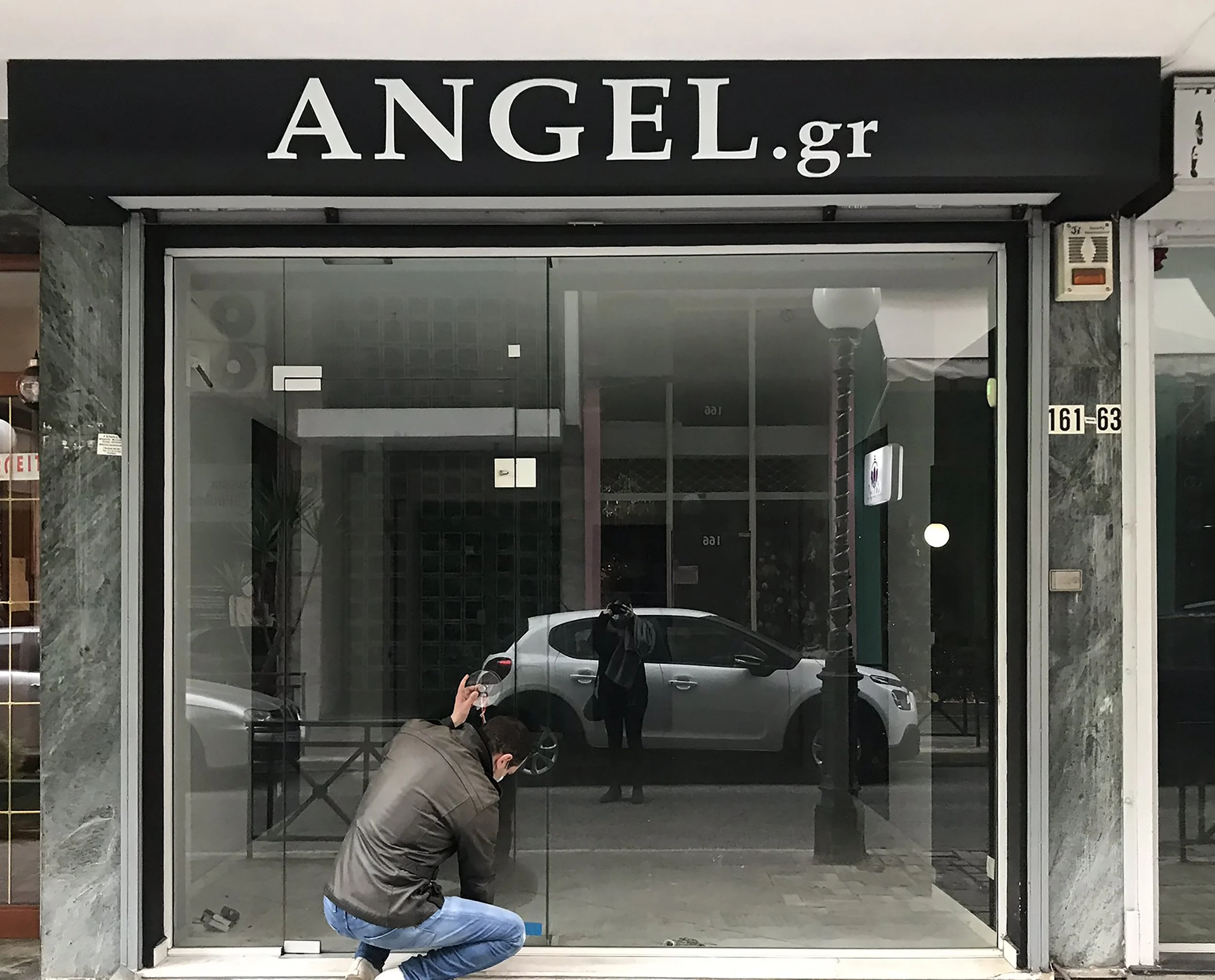 Reflection of street scene with cars, lamppost, and person holding wine glass reflected in storefront window with "ANGEL.gr" sign above.