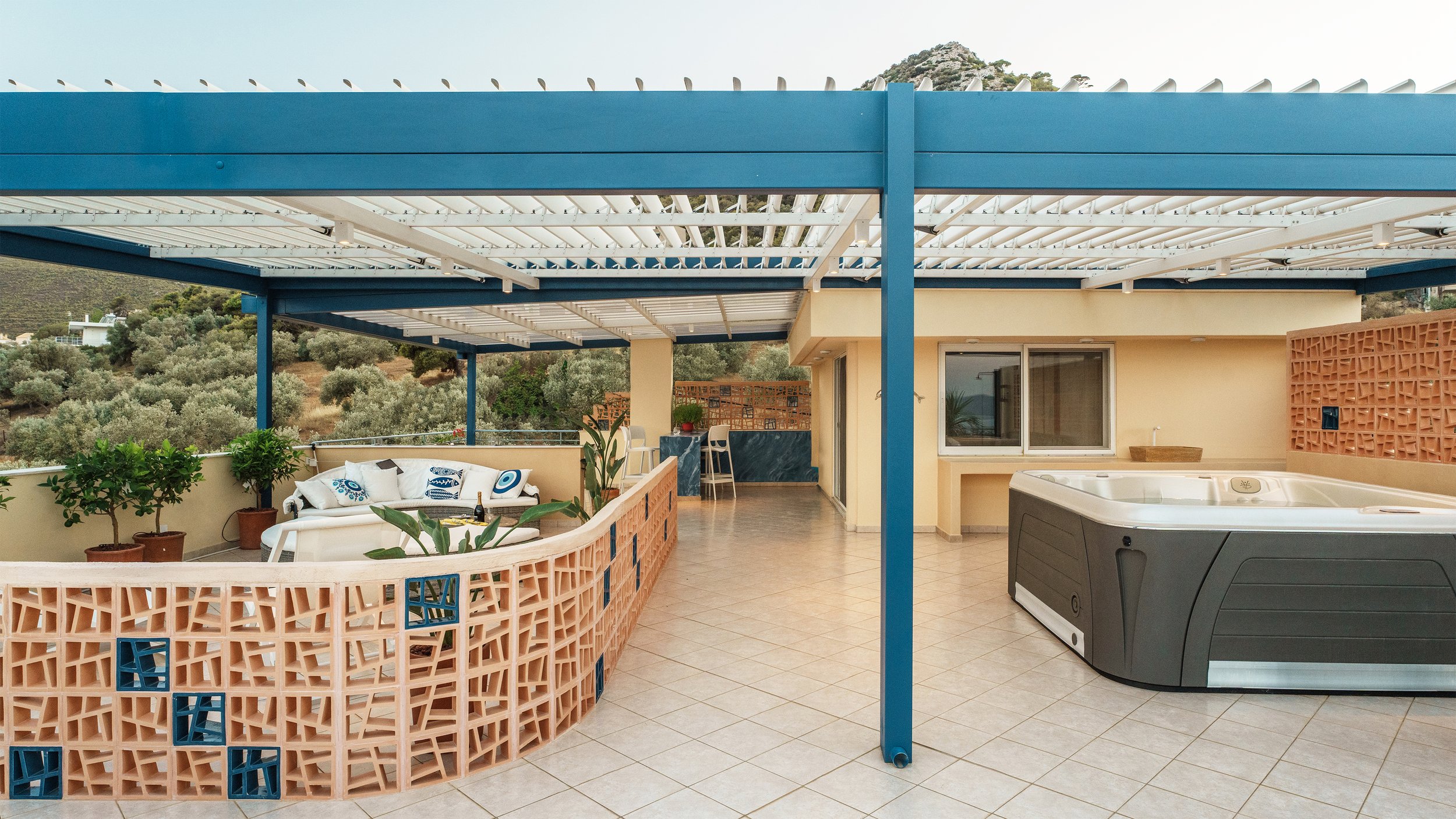 Outdoor terrace with seating area, potted plants, a hot tub, and a view of surrounding greenery and hills. The terrace has a shaded pergola and colorful lattice wall.