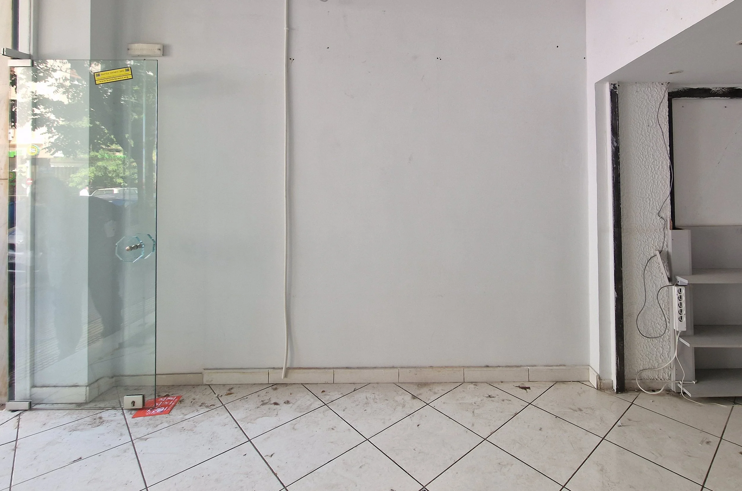 Empty interior space with white walls, a glass door to the left, tiled floor, and visible electrical wiring and outlet.
