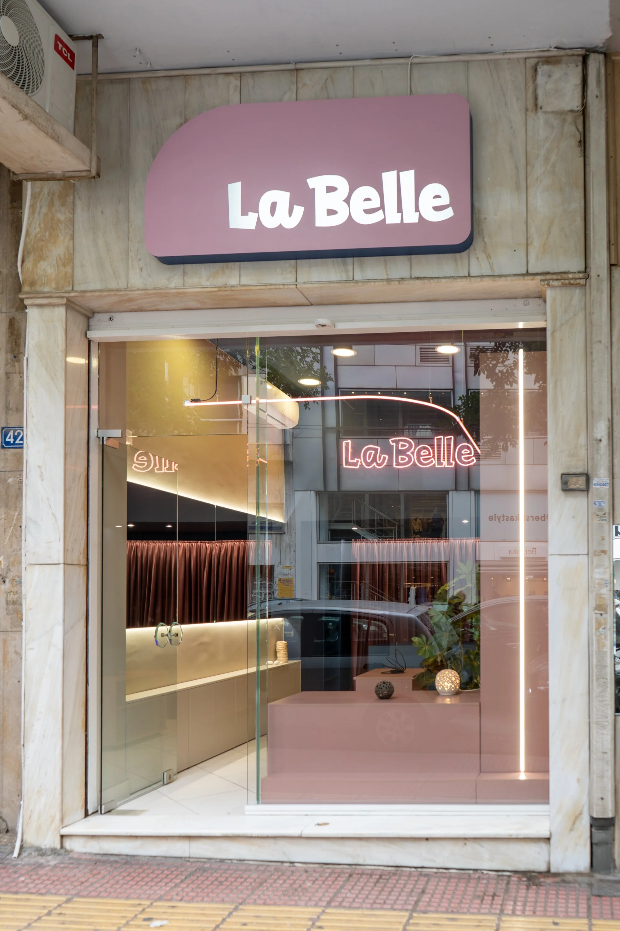 The storefront of La Belle, a jewelry store, with a pink and white sign above the entrance, glass display window showing jewelry and interior decor, and neon signs inside.