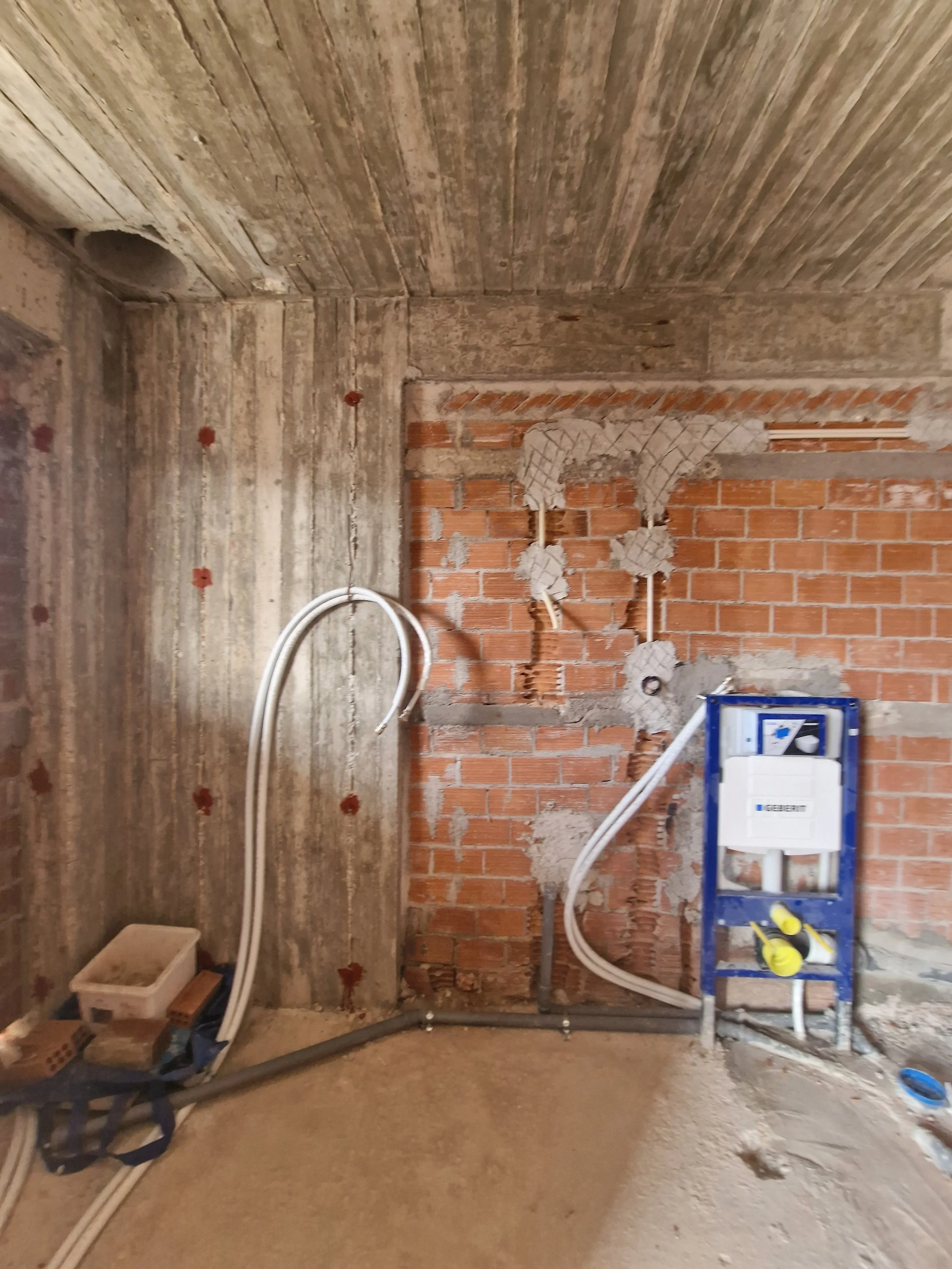 Underside of a building under construction, showing exposed brick wall, plumbing pipes, and electrical wiring, with construction tools and materials on the floor.