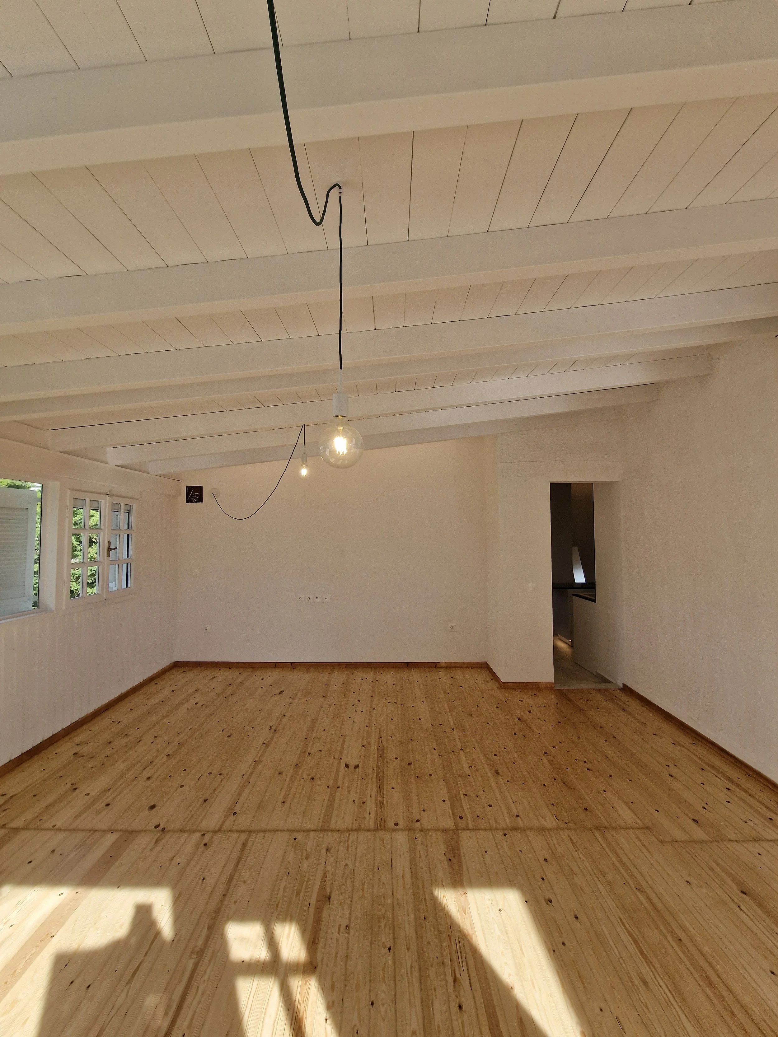 Empty room with wooden floor, white walls, ceiling with exposed beams, two hanging light bulbs, multiple windows, and a small doorway leading to another area.